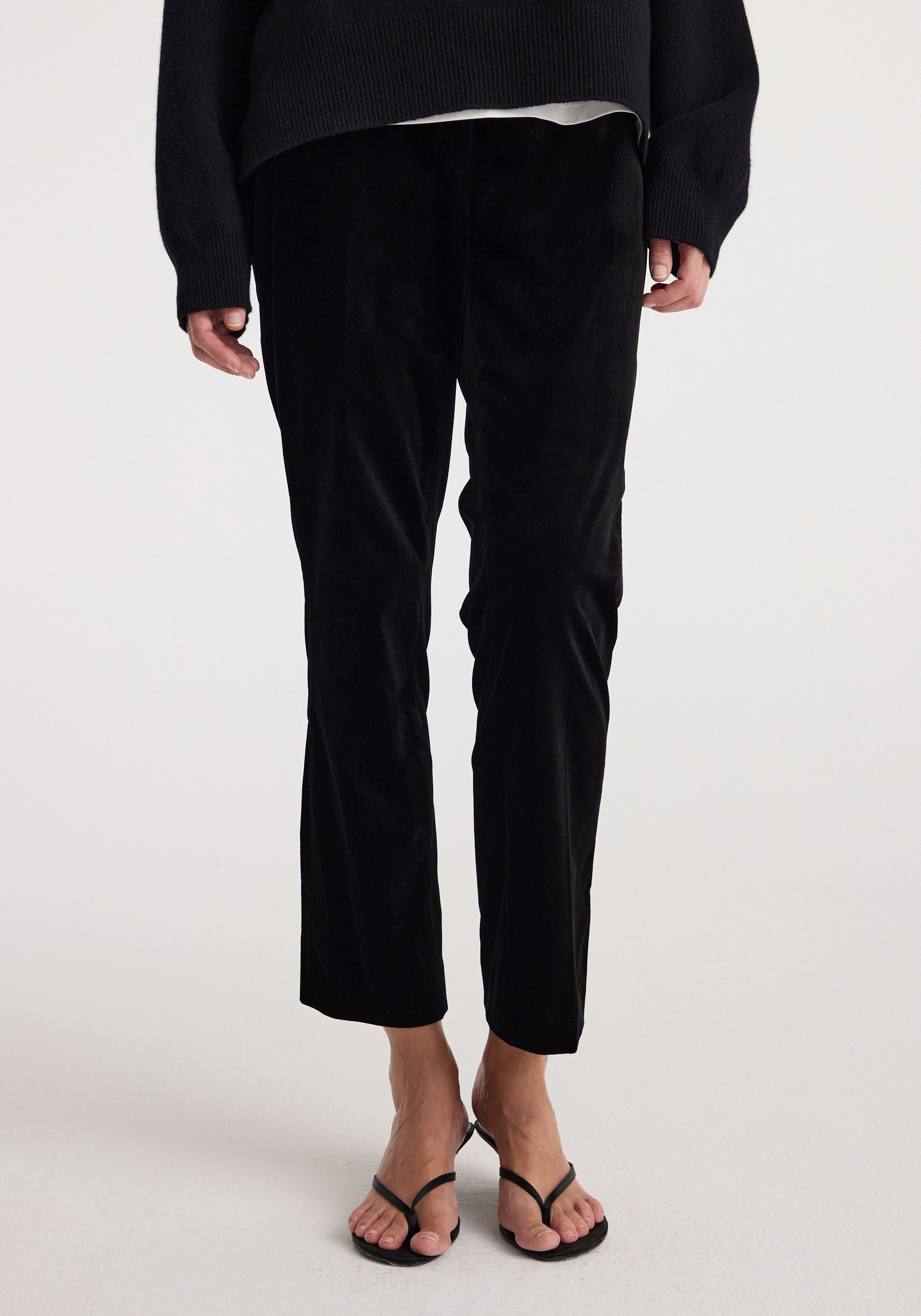 Black cropped trousers worn by a model, paired with a black sweater and black thong sandals, photographed against a clean white studio background.
