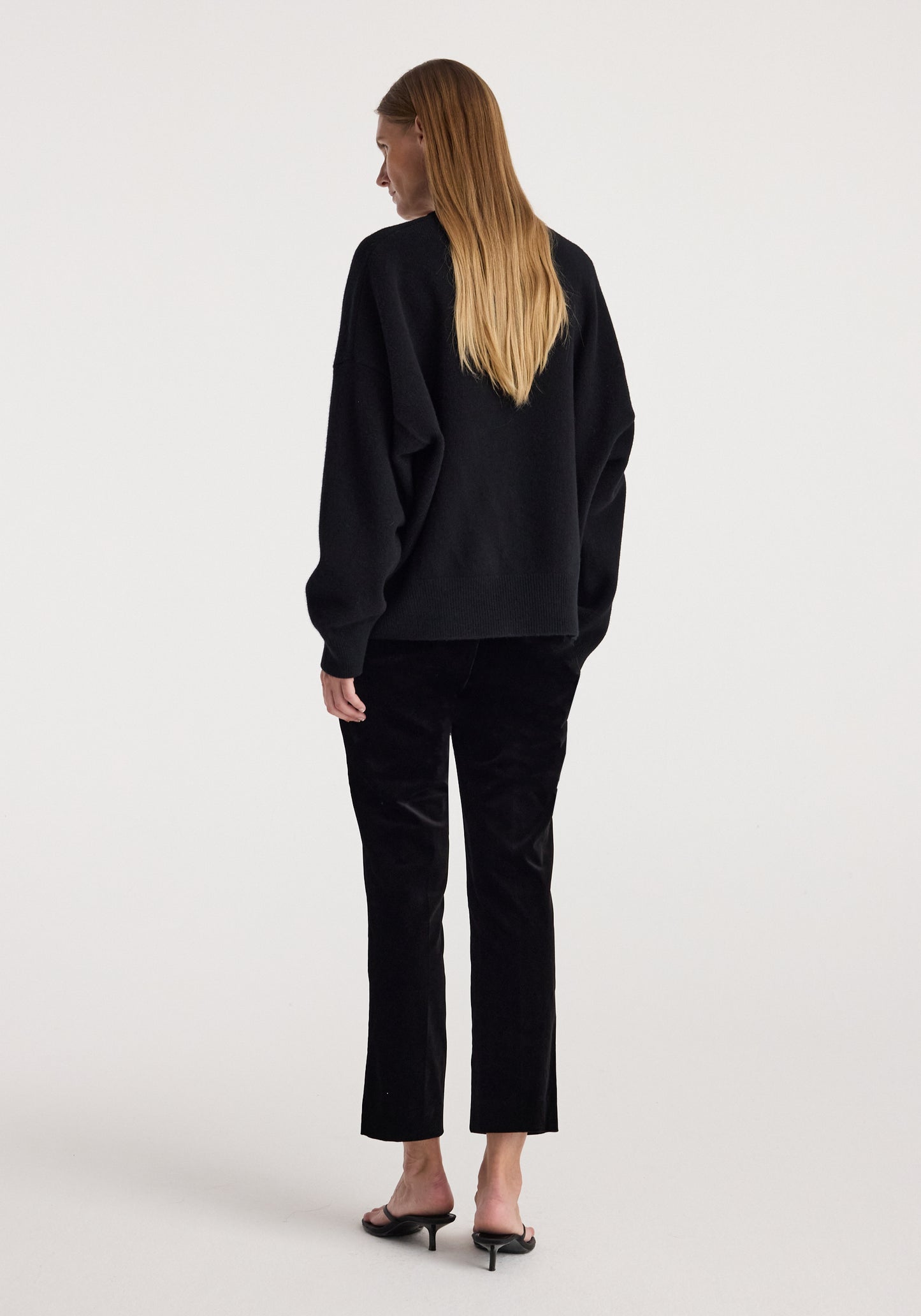 A woman with long blonde hair is shown from the back wearing a black long-sleeve top and black cropped trousers, paired with black heeled mules, against a plain light gray studio background.