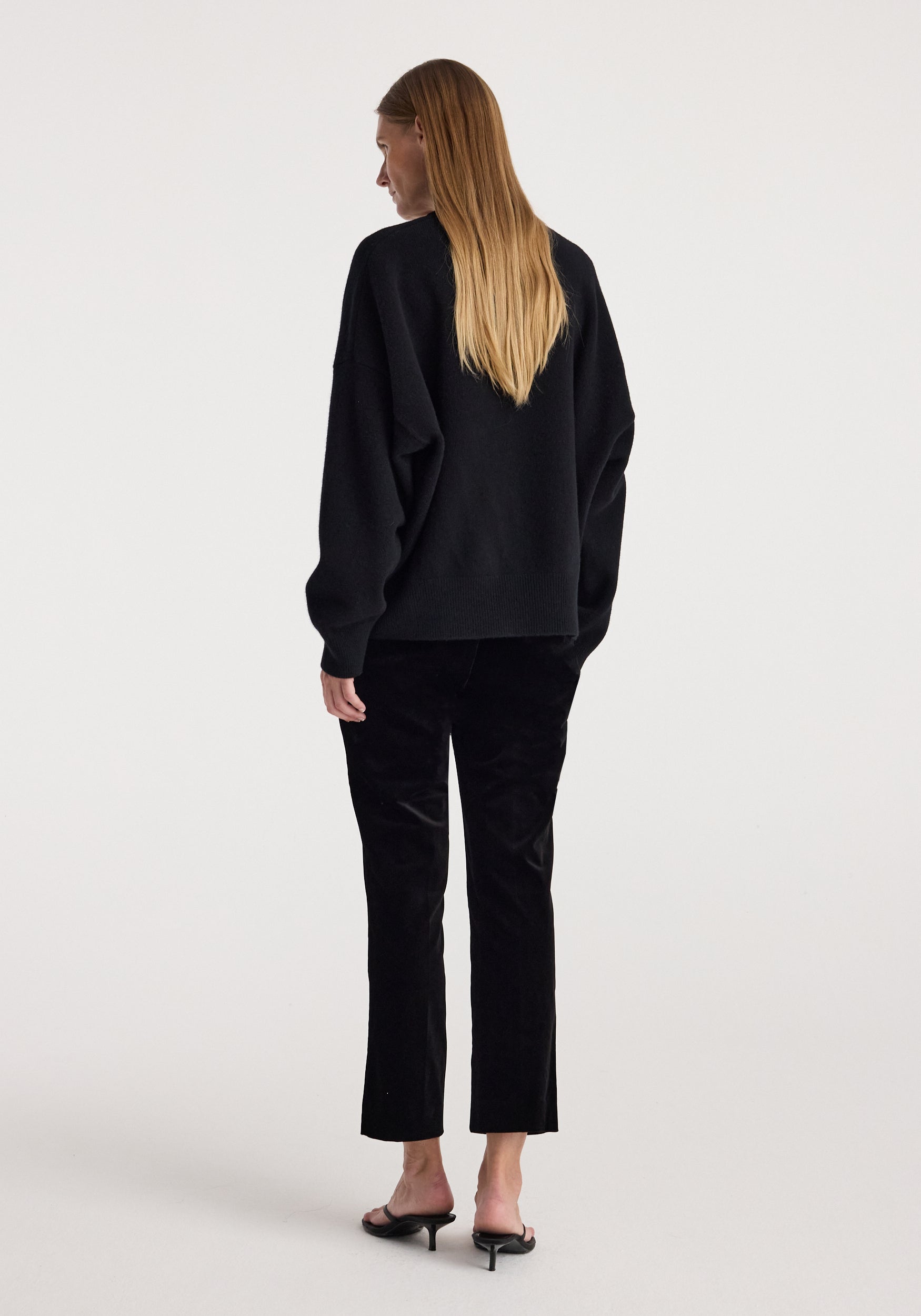 A woman with long blonde hair is shown from the back wearing a black long-sleeve top and black cropped trousers, paired with black heeled mules, against a plain light gray studio background.