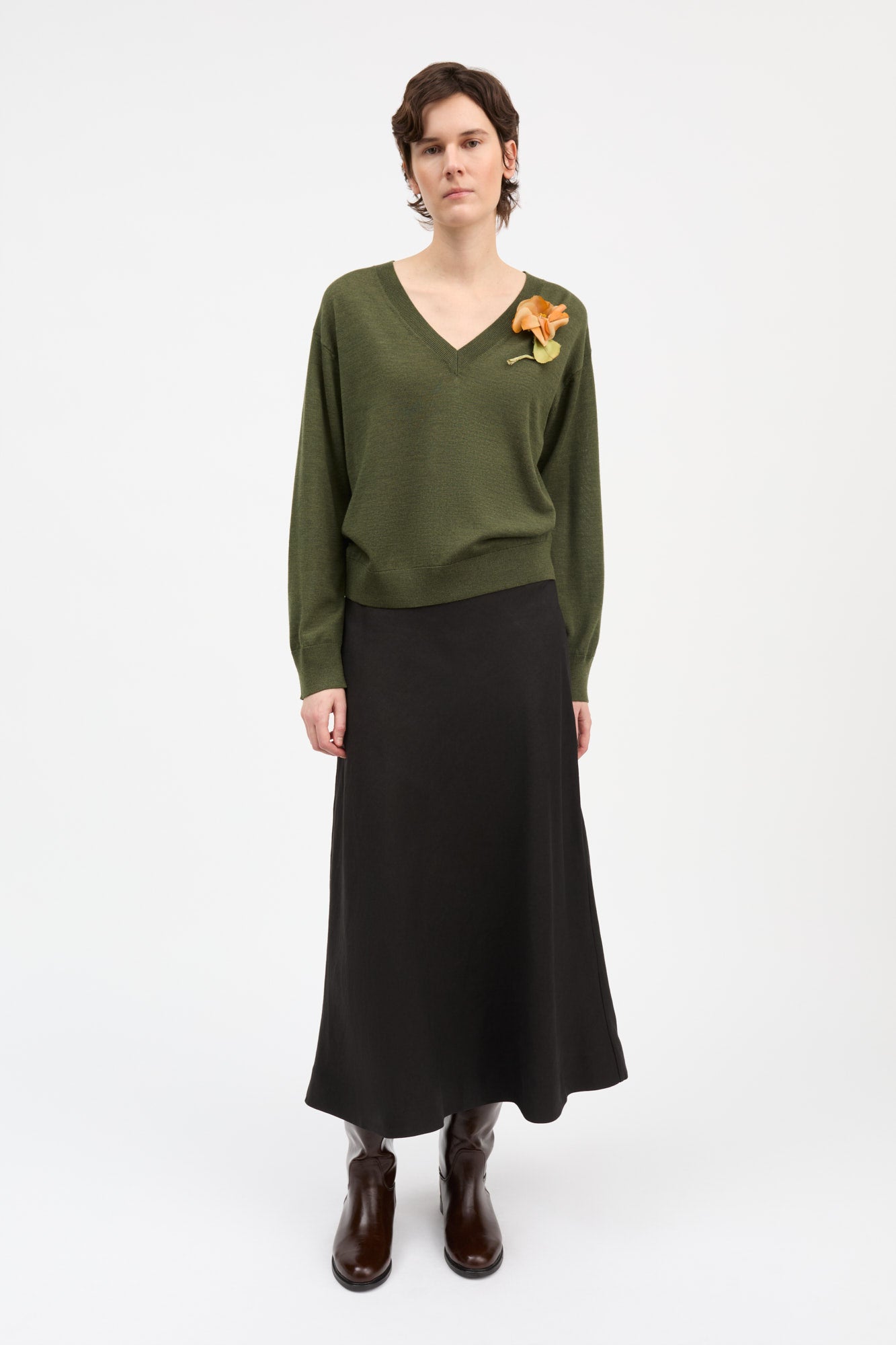 A model stands against a plain white studio background wearing a dark olive green long-sleeve knit sweater with a yellow-orange flower applique on the chest, paired with a long black skirt and dark brown ankle boots.