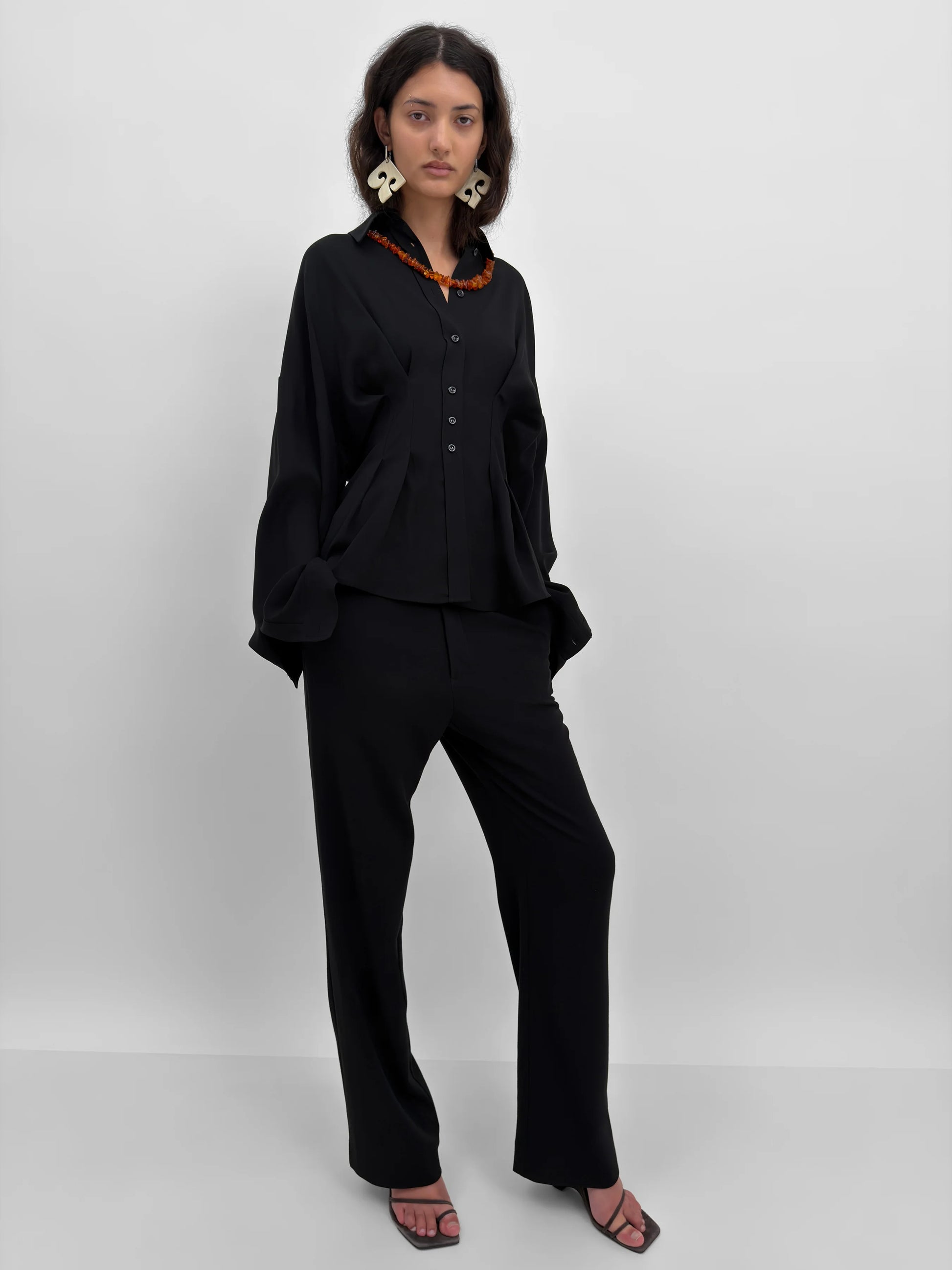 A model wearing a black long-sleeve button-front top with a wide collar and matching black wide-leg trousers, styled with a chunky orange necklace and large white earrings against a light gray studio background.