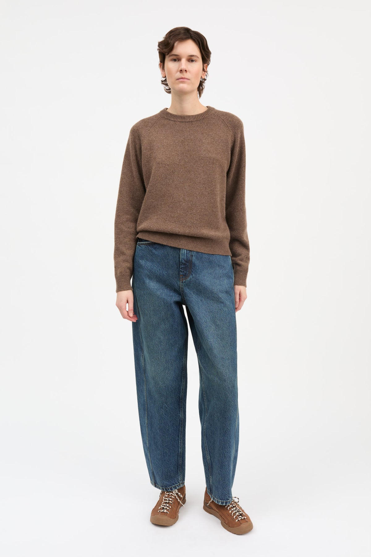 A model stands against a plain white studio background wearing a brown crewneck knit sweater, blue wide-leg jeans, and brown lace-up shoes.