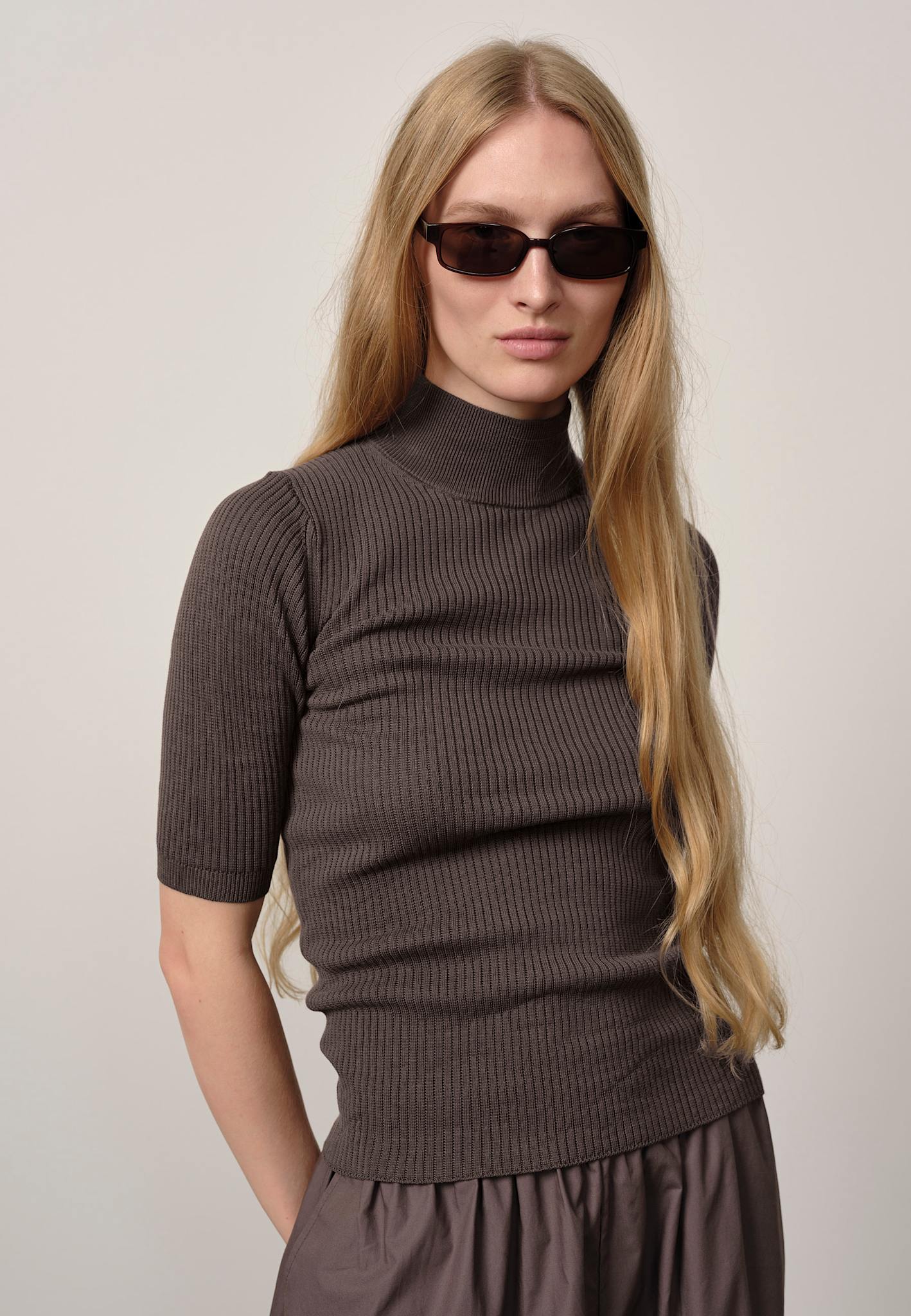 Woman wearing a brown ribbed turtleneck sweater and sunglasses against a plain background