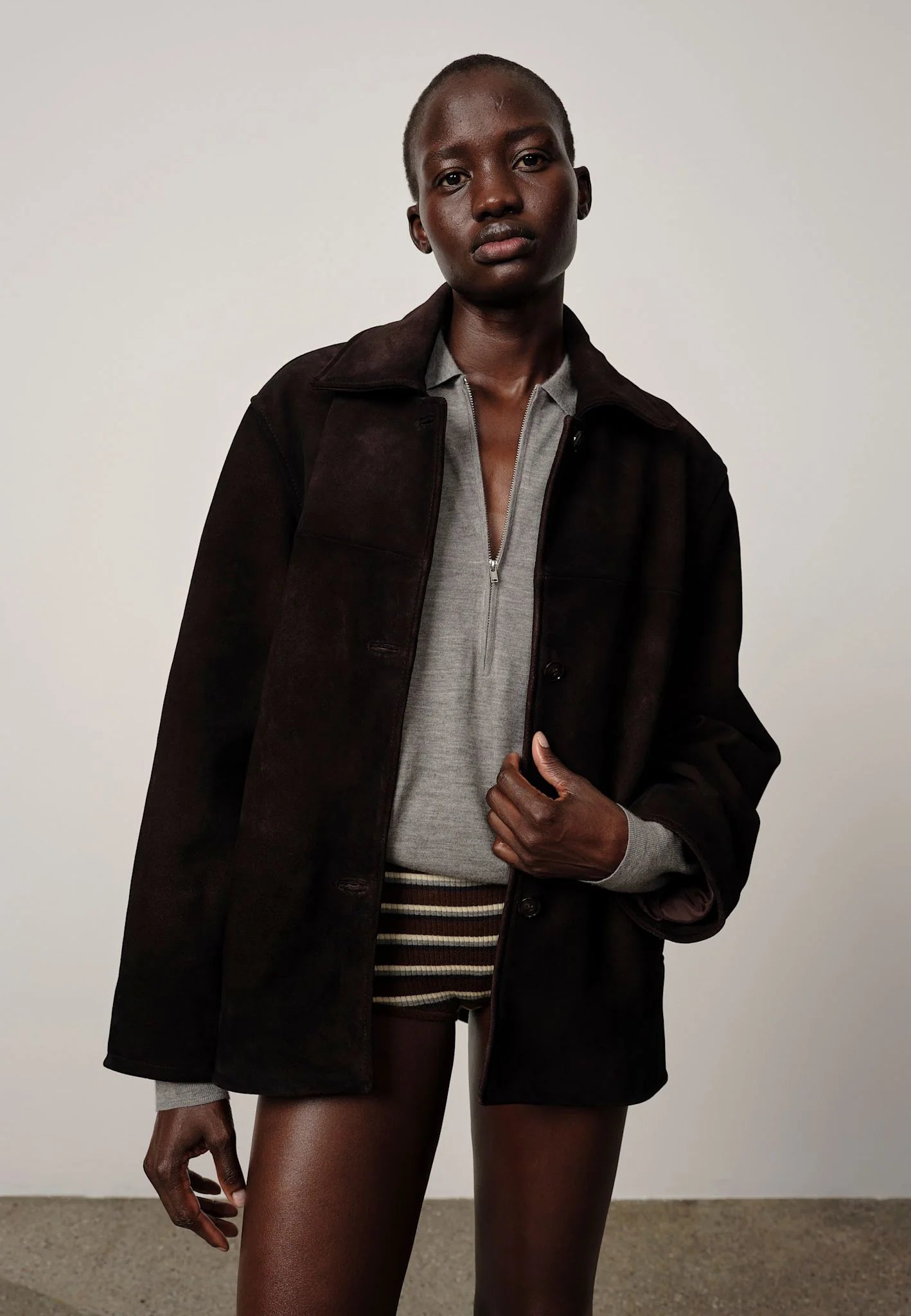 Person wearing a dark coat over a light shirt and striped shorts on a plain background