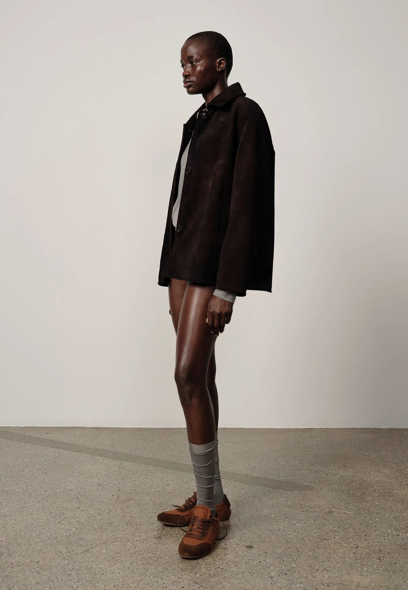 Person wearing a dark jacket, shorts, and brown shoes on a plain background