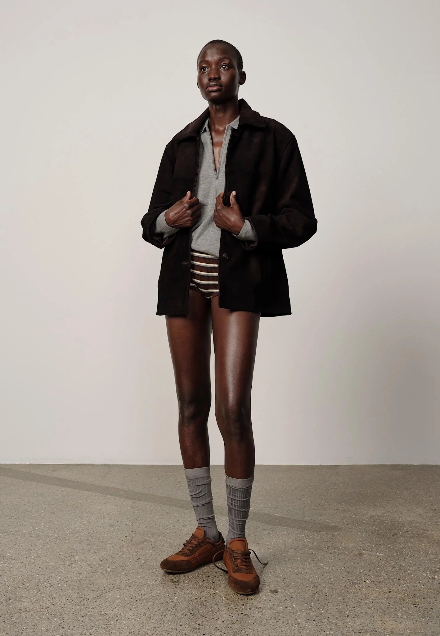 A model stands in a studio wearing a black overshirt over a light gray shirt, brown and white striped shorts, gray socks, and brown shoes against a plain off-white wall and concrete floor.