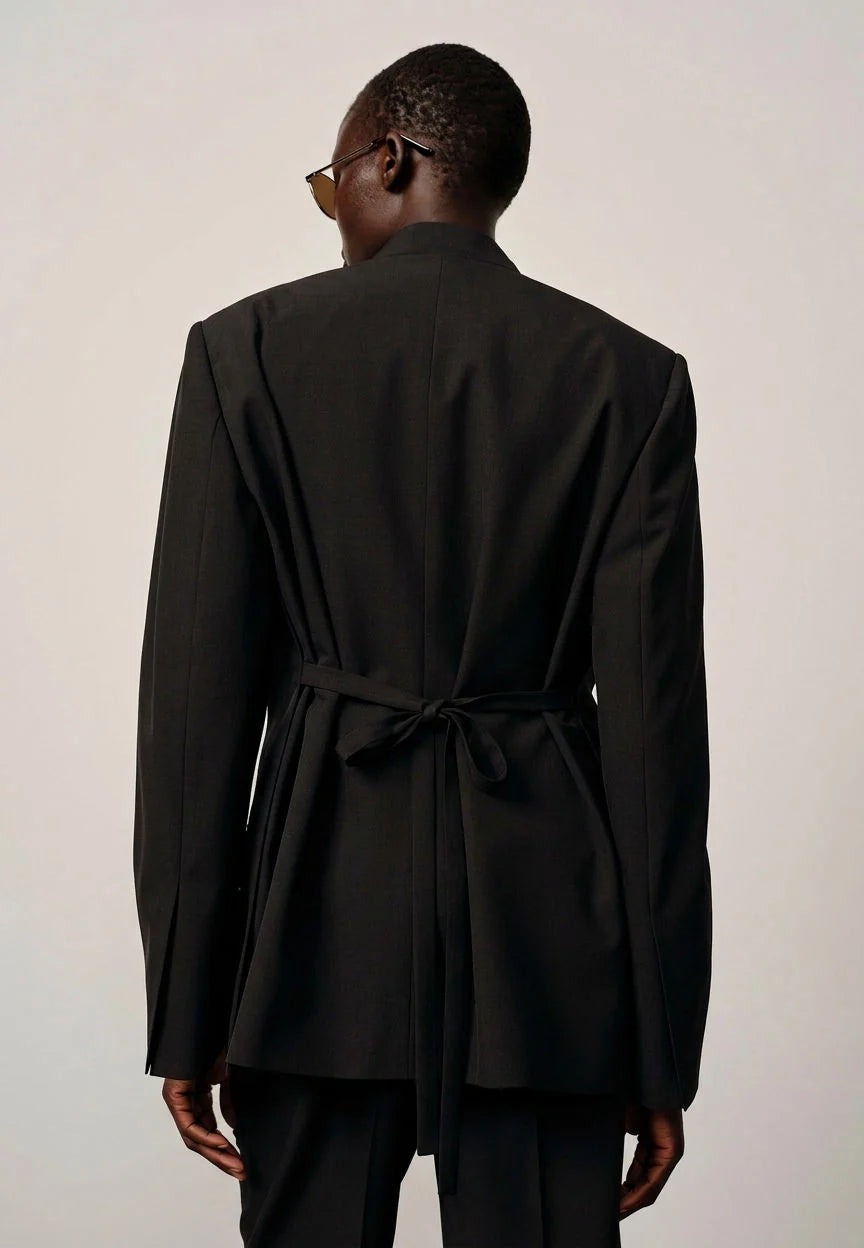 Person wearing a black blazer with a tie waist on a plain background