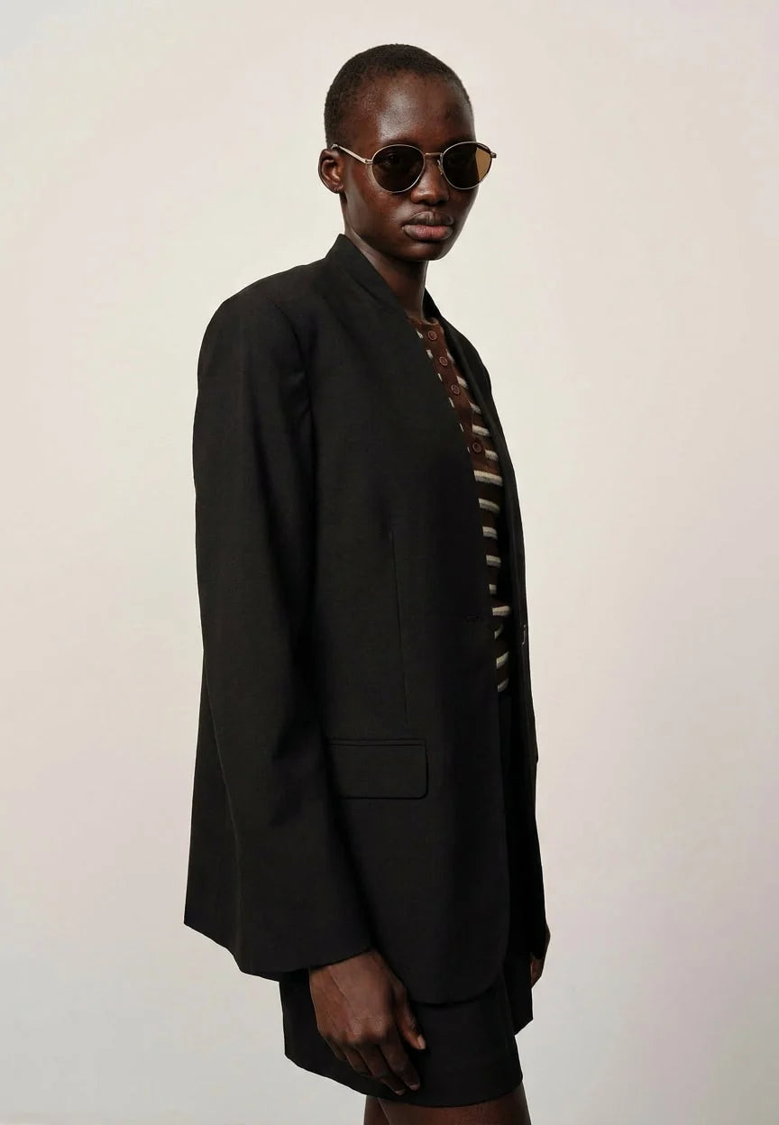 A model wearing a black oversized blazer and matching shorts over a brown and white striped shirt, styled with round sunglasses, against a plain light background.