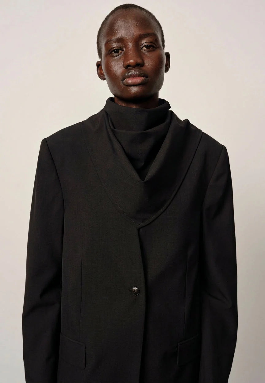 A model wearing a black cowl-neck blazer with a matching black turtleneck, photographed against a plain light gray background.