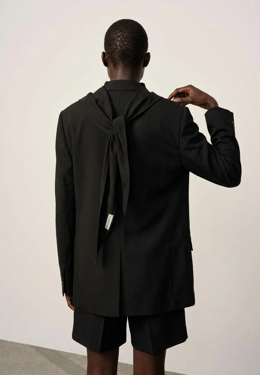 Person wearing a black coat with a bow detail on the back, standing against a plain white wall.