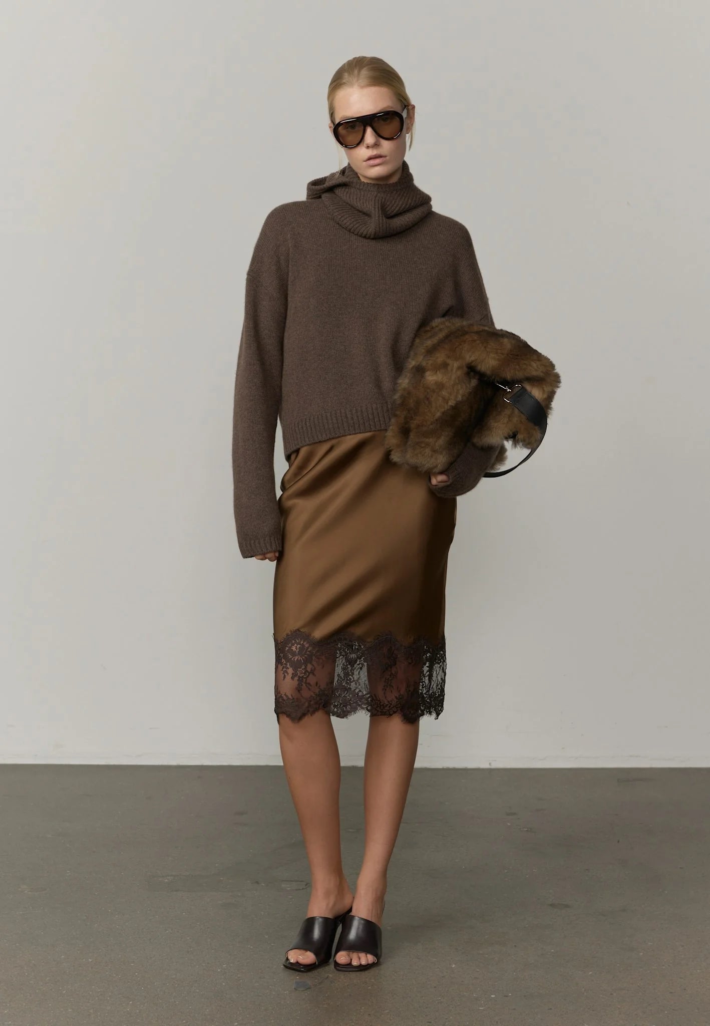 A model wearing a brown knit sweater and a brown satin pencil skirt with a black lace hem, paired with black slide sandals.