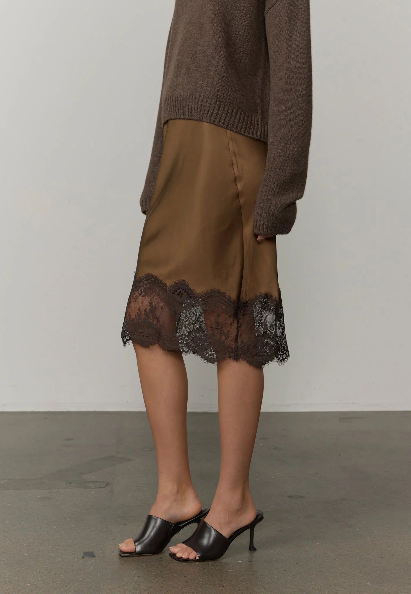 A model is shown from the waist down wearing a brown knit sweater, a brown satin midi skirt with a black lace hem, and black leather slide heels against a neutral studio background.