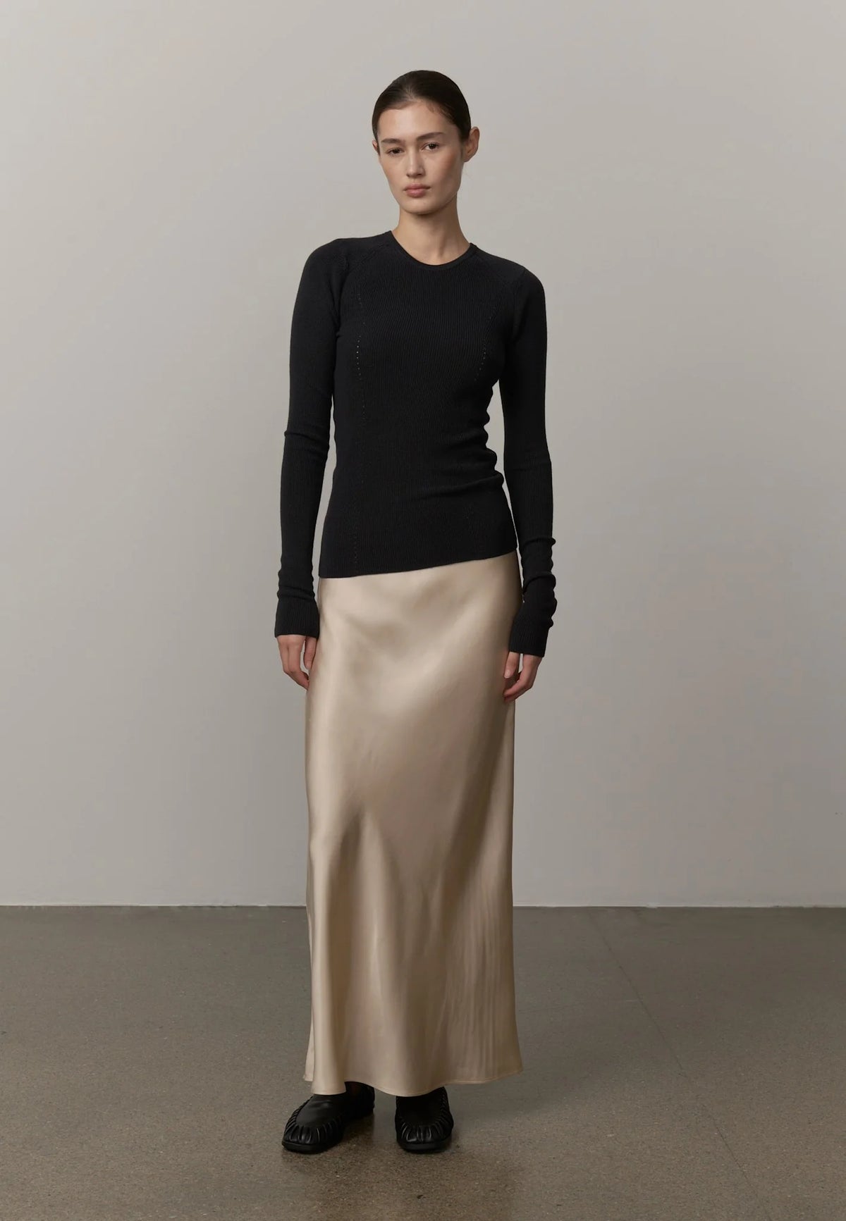 Woman wearing a black long-sleeve top and beige satin skirt against a neutral background