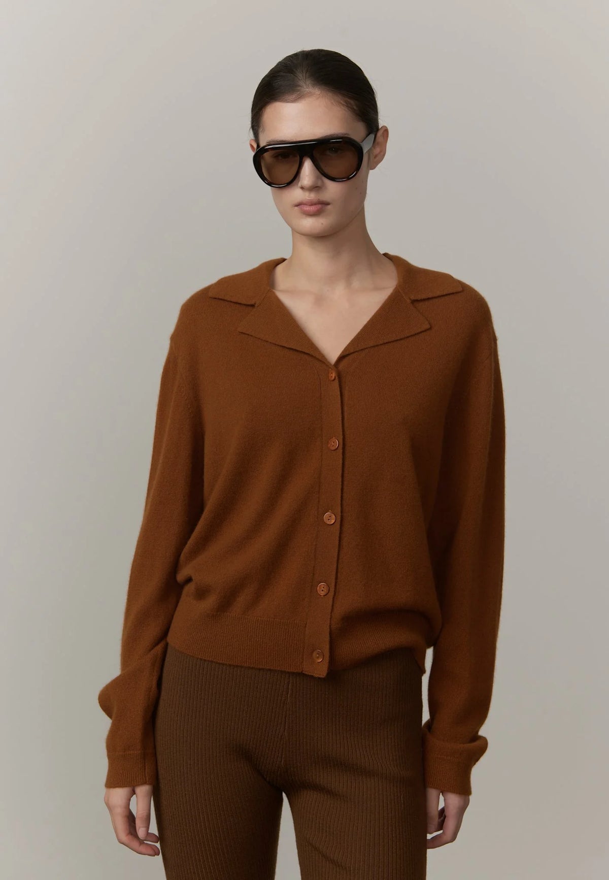 A model wearing a rust-brown knit button-front cardigan with a collar and matching brown ribbed trousers, styled with oversized black sunglasses, against a plain light gray studio background.