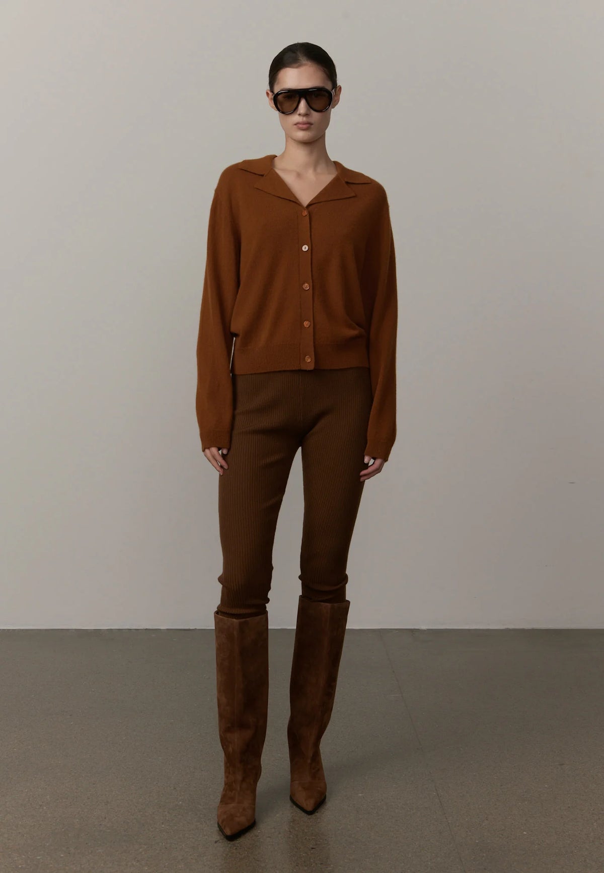 A model stands in a studio wearing a matching brown knit cardigan and slim-fit pants, paired with tall brown suede boots and dark sunglasses, against a neutral light gray background.