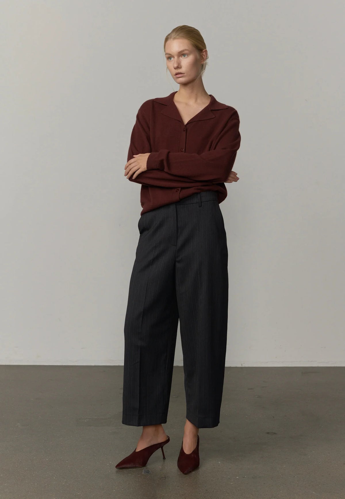 A woman stands in a studio wearing a burgundy long-sleeve collared knit top, dark charcoal cropped trousers, and burgundy pointed-toe heels against a plain light gray wall and concrete floor.