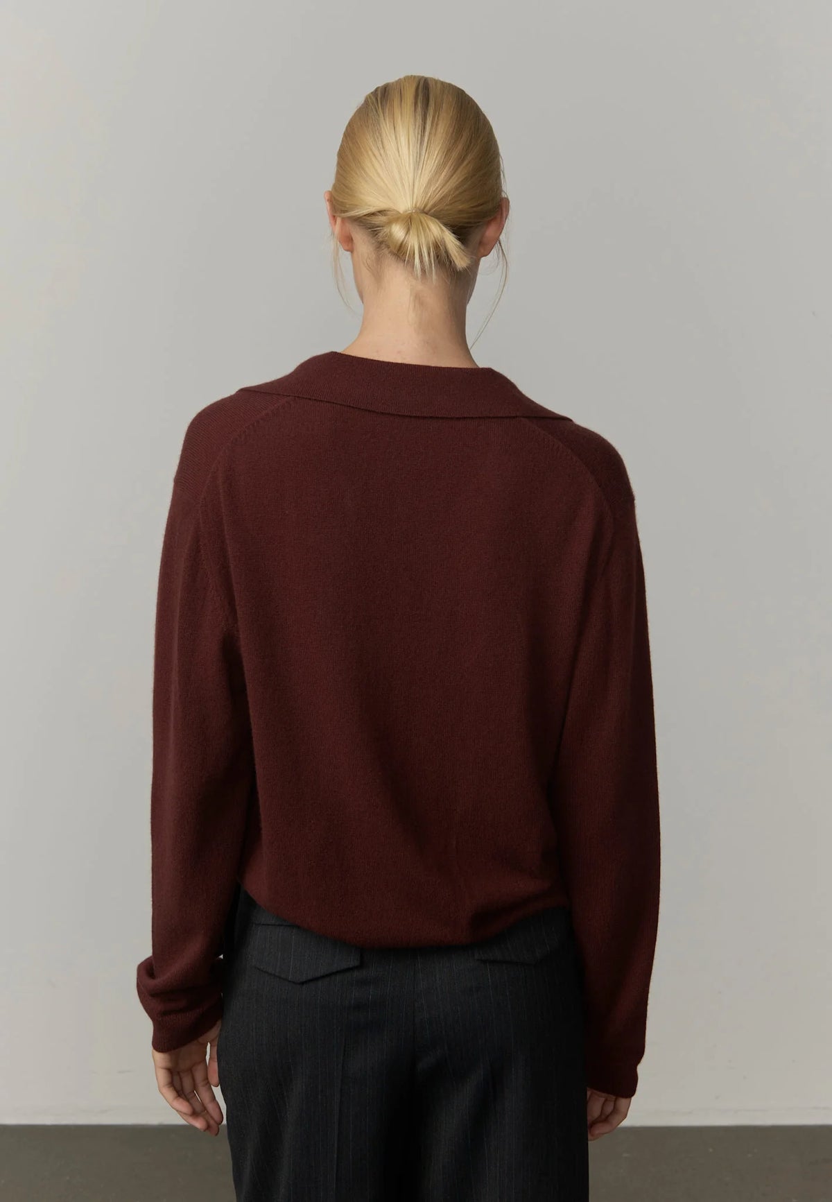 A woman viewed from behind wearing a dark burgundy long-sleeve knit top and dark trousers, standing against a plain light gray wall.