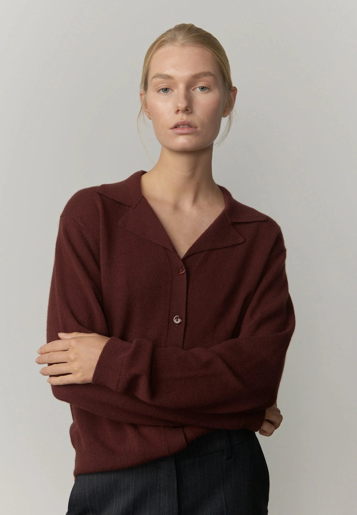 A blonde model wearing a dark burgundy knit cardigan with a collar and button detail, paired with dark trousers, against a plain light gray studio background.