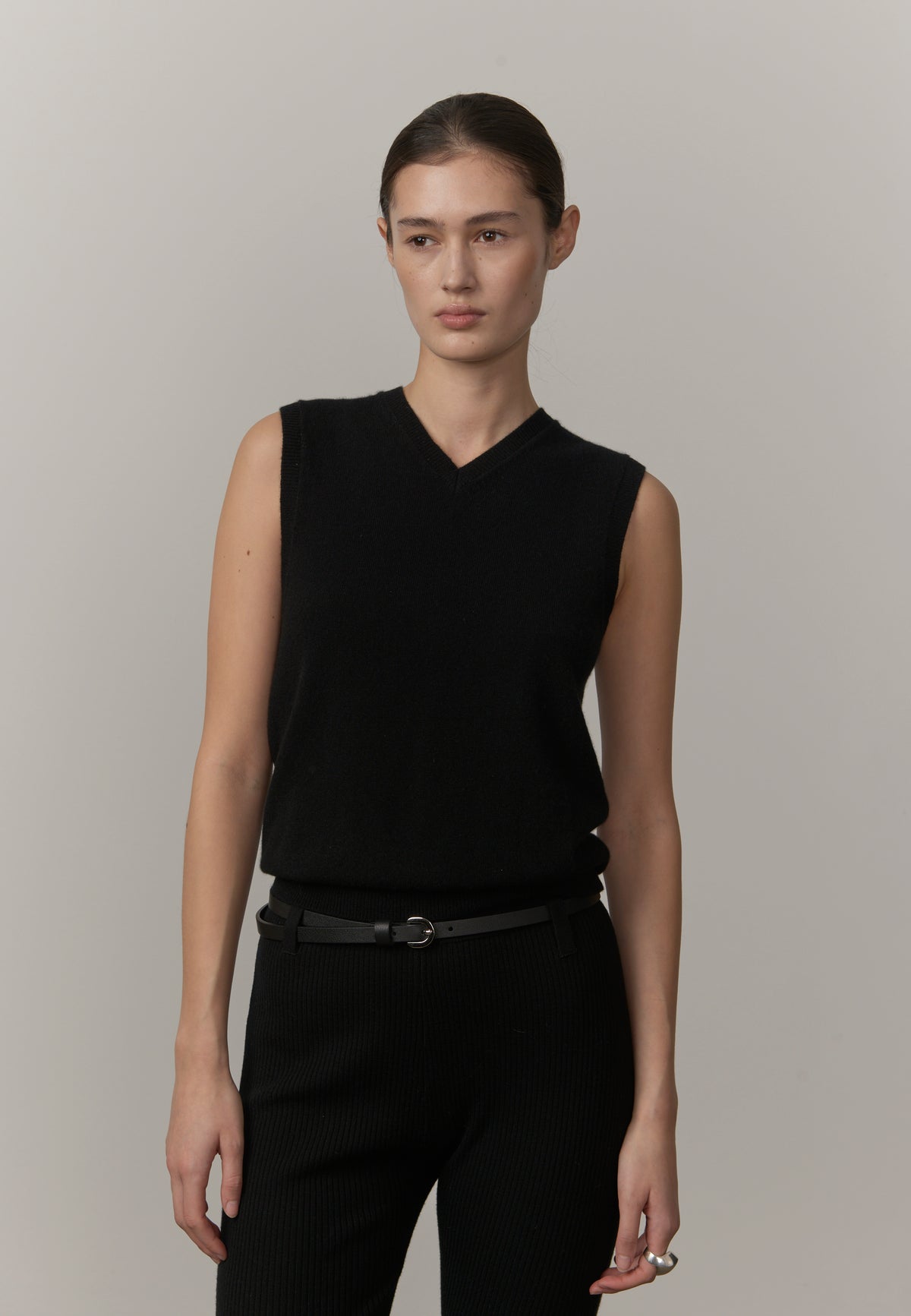 A model wearing a black sleeveless knit V-neck sweater vest paired with black trousers and a black belt, standing against a neutral light gray studio background.