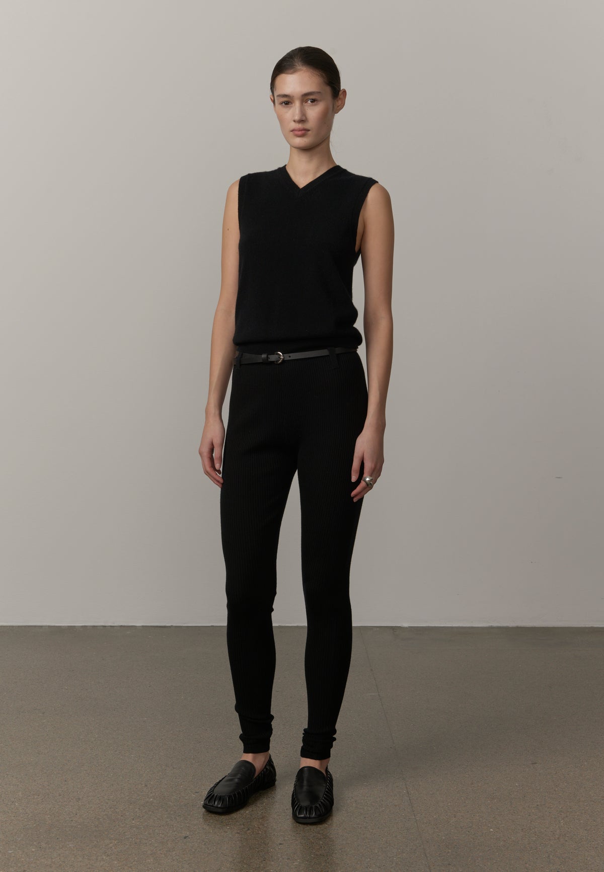 A model stands in a studio wearing a black sleeveless V-neck knit top, black pants, a thin black belt, and black loafers against a plain light gray wall and concrete floor.