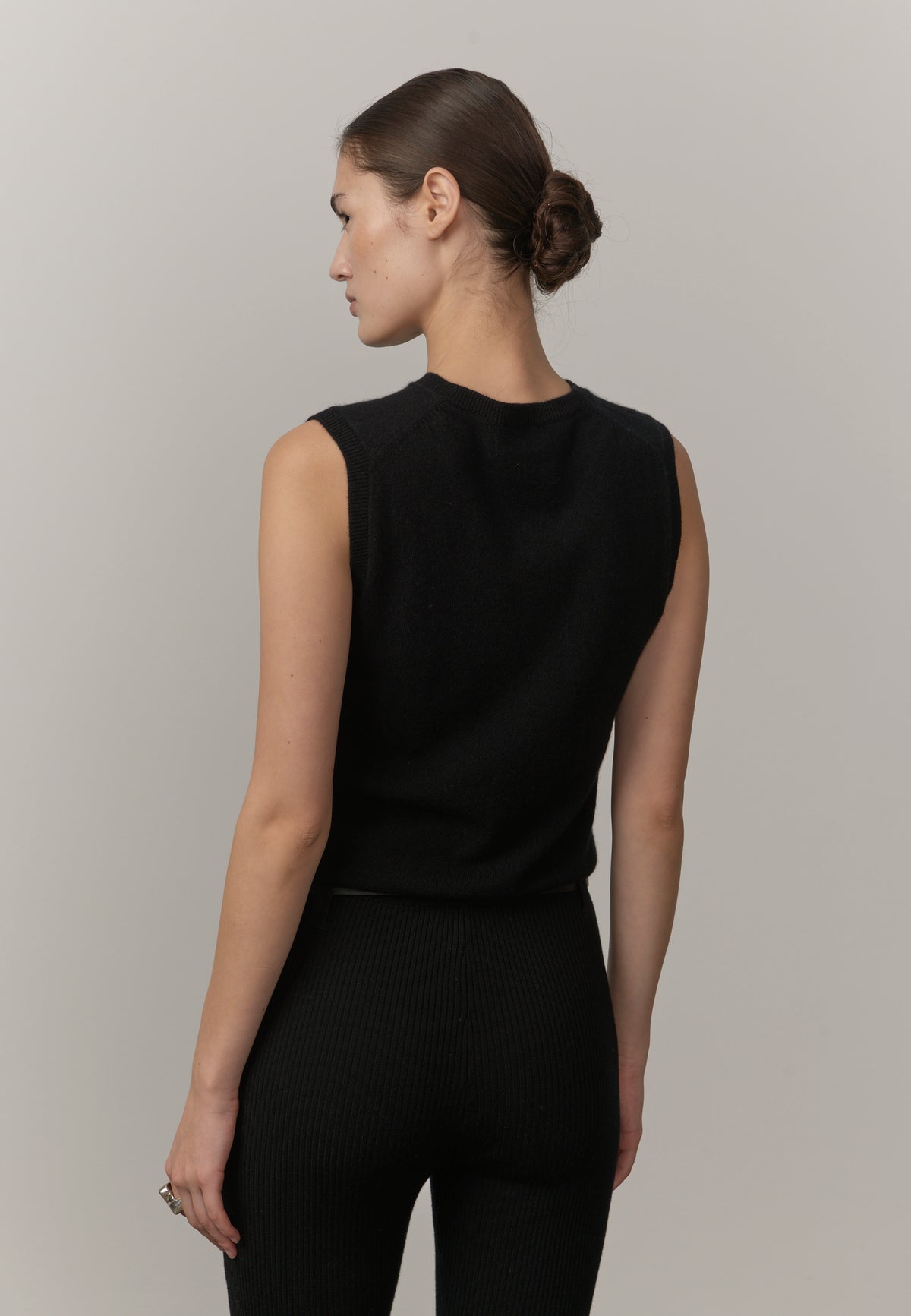 A woman shown from the back wearing a sleeveless black ribbed knit top and matching black ribbed leggings against a plain light gray studio background.