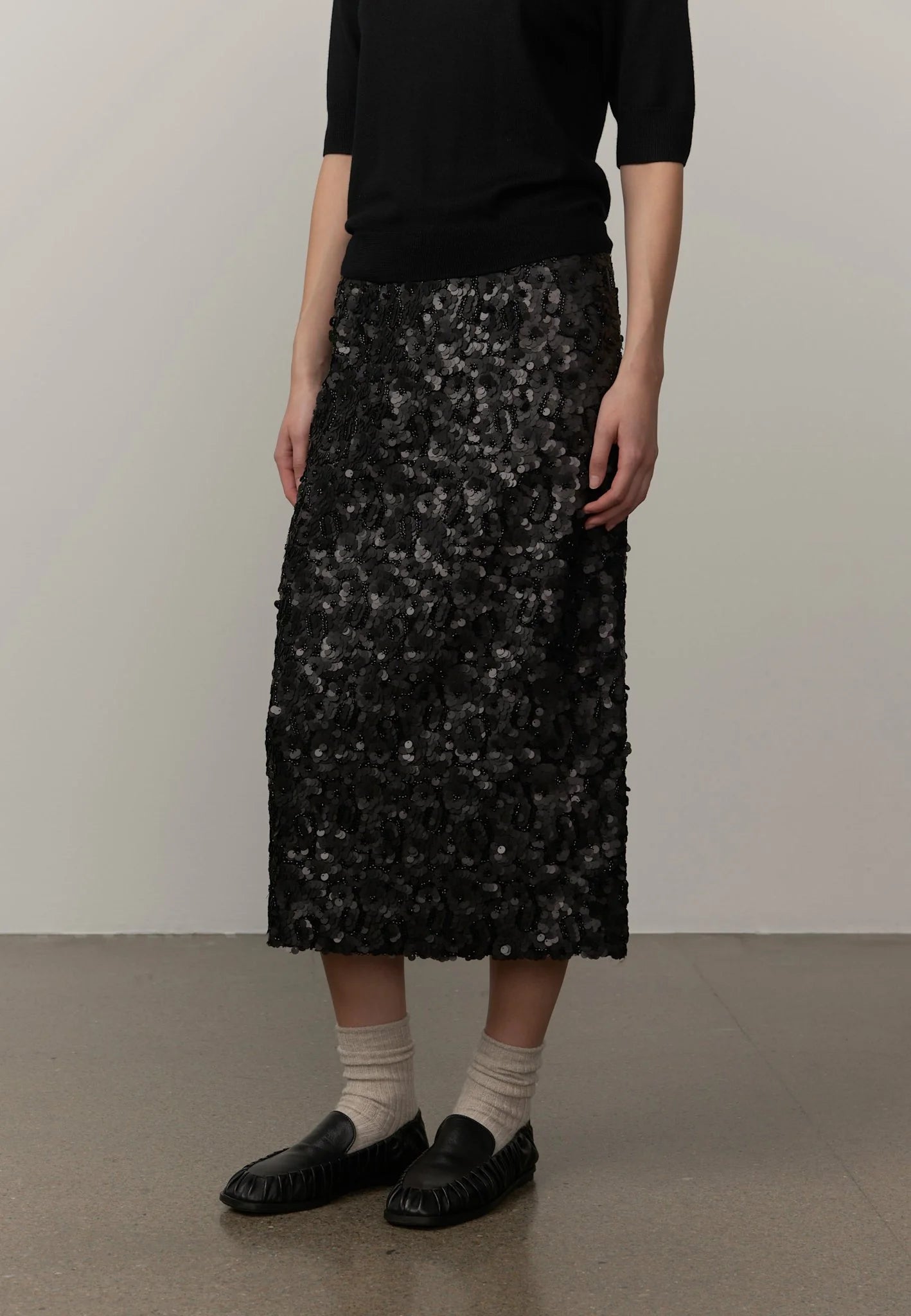 A model wearing a black midi skirt covered in textured, scale-like black and gray sequins or beads, paired with a black top, beige socks, and black loafers, standing against a plain light gray wall.
