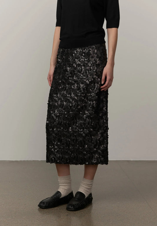 A model wearing a black midi skirt covered in textured, scale-like black and gray sequins or beads, paired with a black top, beige socks, and black loafers, standing against a plain light gray wall.