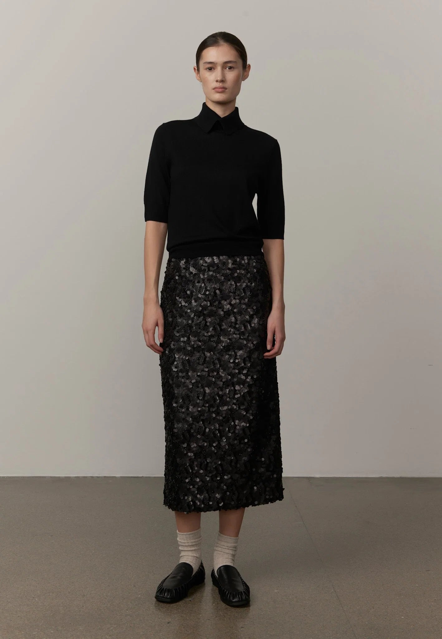 A model stands in a studio wearing a black short-sleeve collared knit top and a black floral lace midi skirt, paired with cream socks and black loafers.