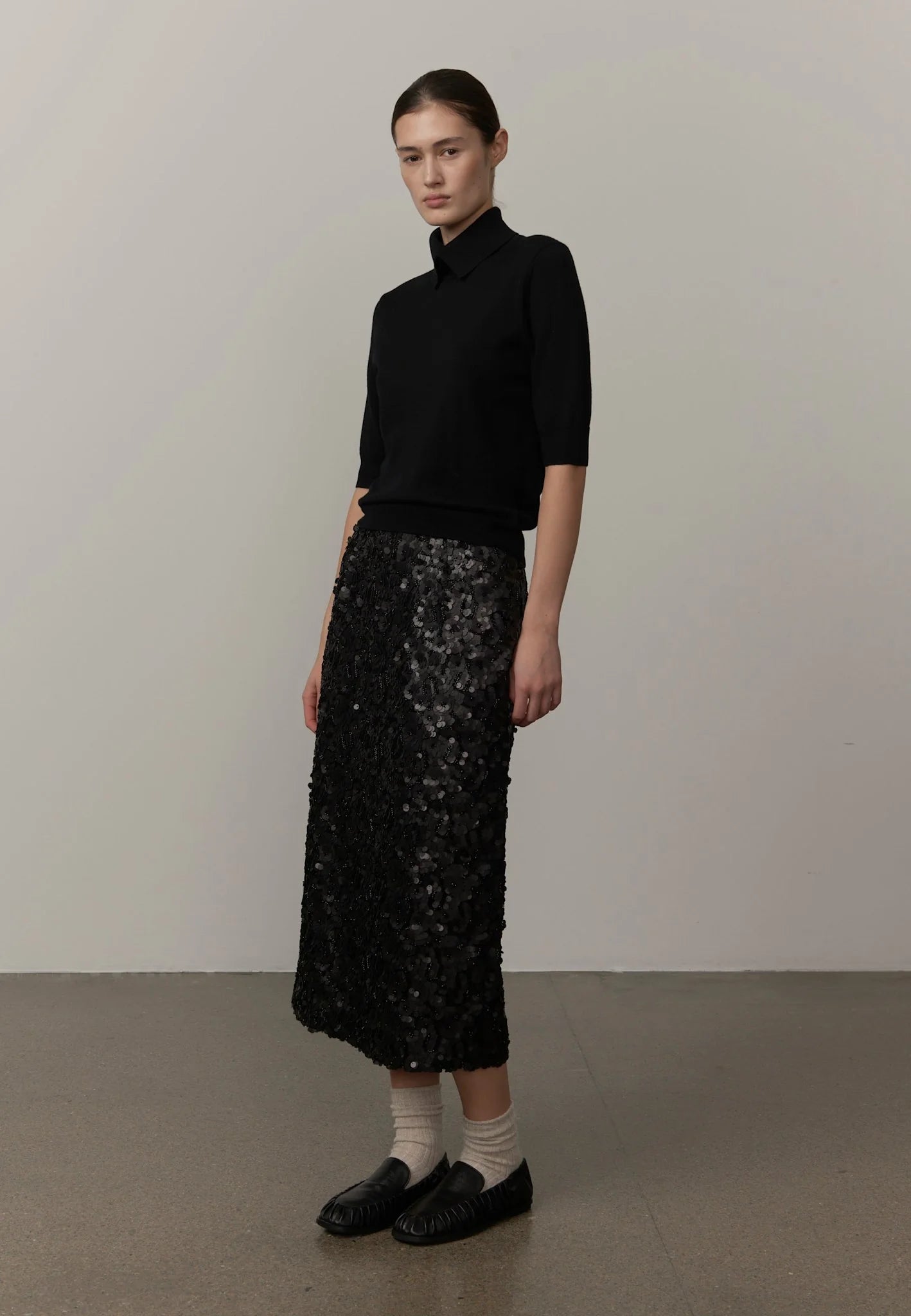 A model stands in a studio wearing a black mock-neck knit top, a black sequined midi skirt, white socks, and black loafers against a plain light gray wall and concrete floor.