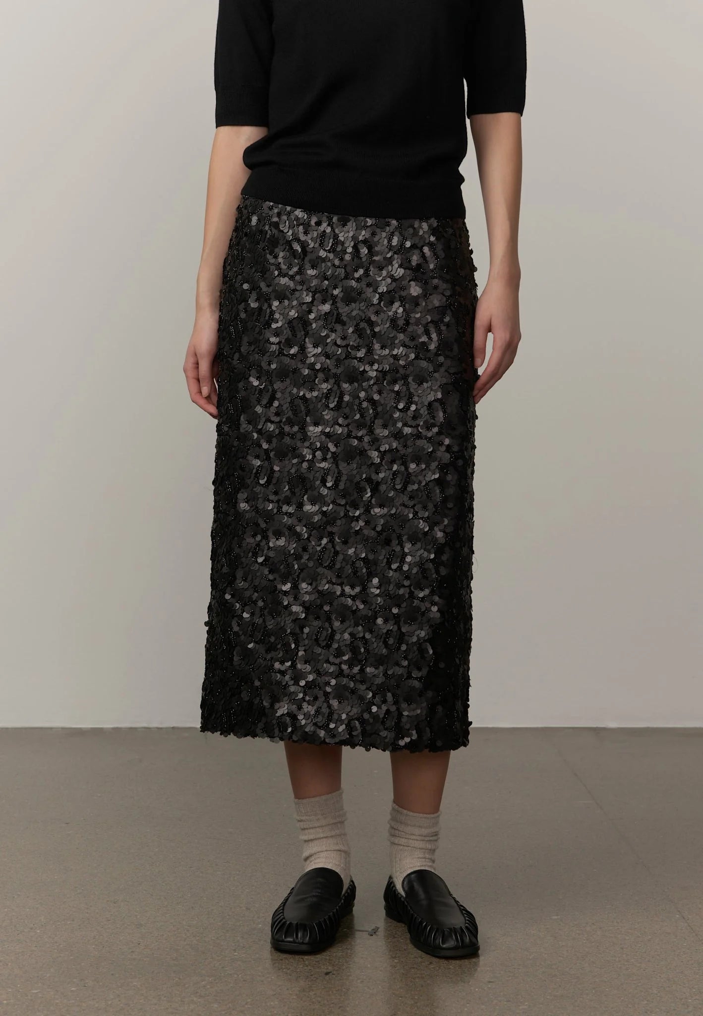 A model wearing a black textured midi skirt with a floral pattern, paired with a black top, beige socks, and black leather loafers, standing against a plain light gray wall.