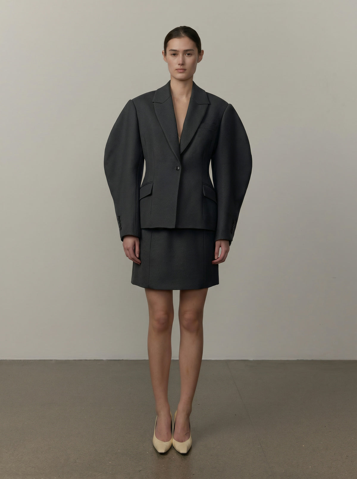 A model stands facing forward in a dark charcoal gray two-piece suit featuring a structured, exaggeratedly voluminous blazer and matching mini skirt, paired with beige pointed-toe heels against a neutral studio backdrop.
