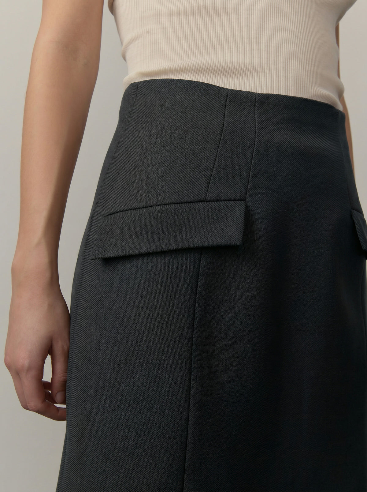 Close-up of a model wearing a dark charcoal gray skirt with a structured waistband and a flap pocket, paired with a ribbed beige top against a neutral gray background.