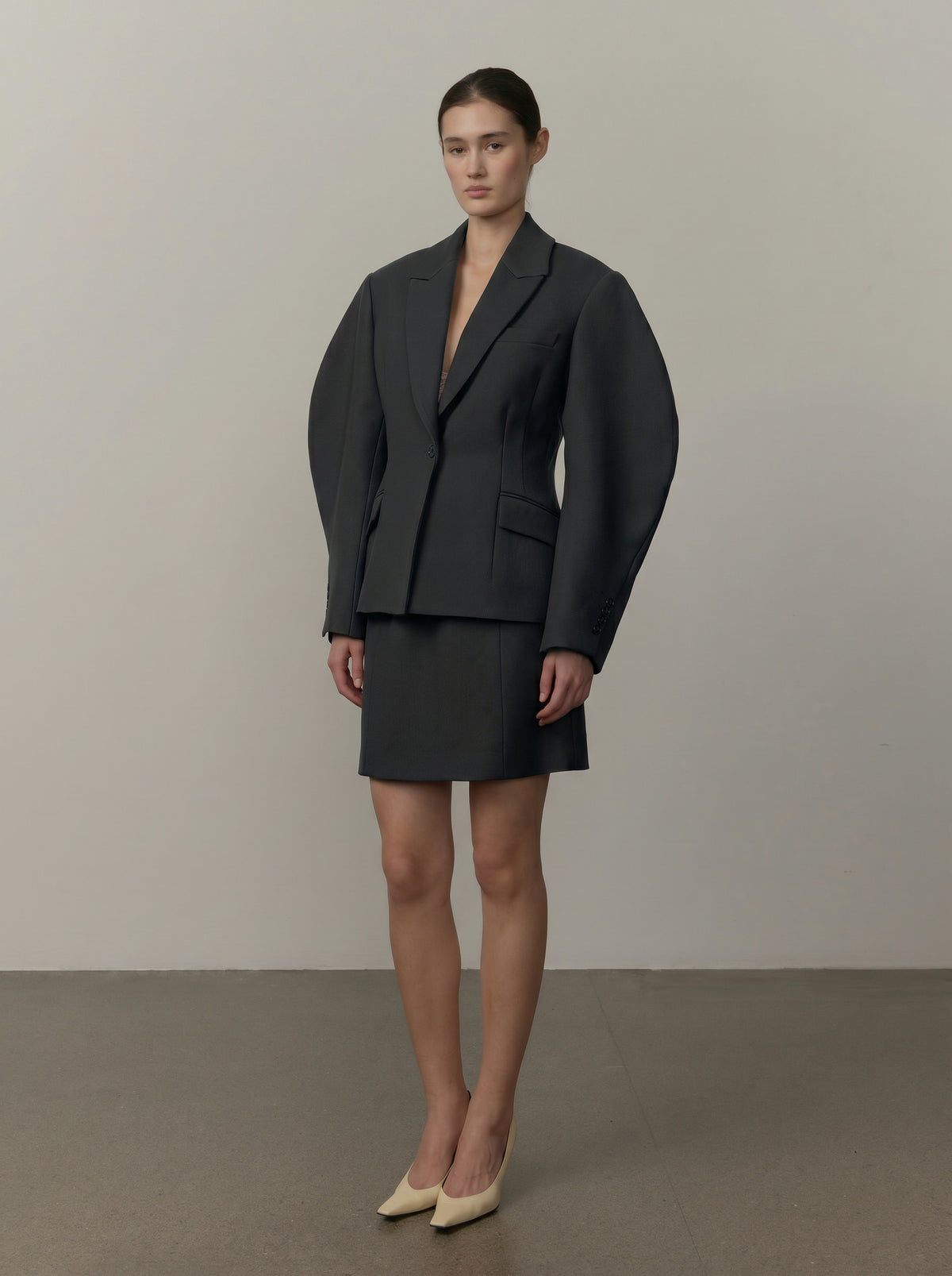 A model stands in a studio wearing a dark charcoal gray two-piece suit featuring a structured blazer with dramatic balloon sleeves and a matching mini skirt, paired with beige pointed-toe heels.