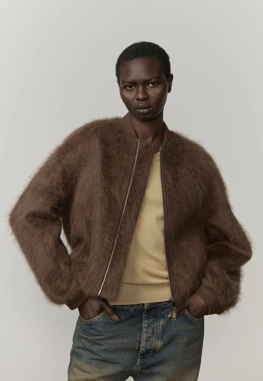 Person wearing a brown fuzzy jacket over a beige shirt and blue jeans on a plain background
