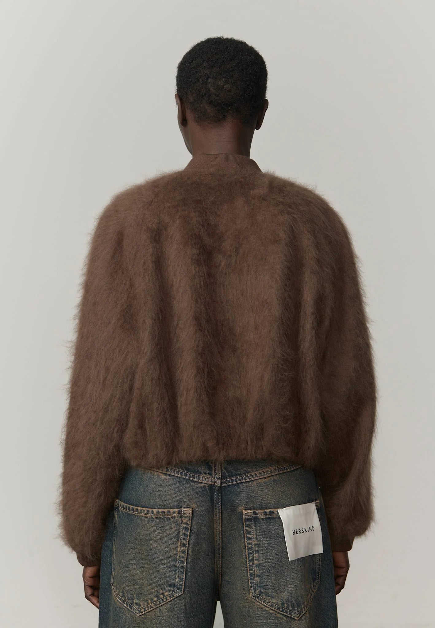 Person wearing a brown fuzzy sweater and blue jeans with a visible brand label on a plain background