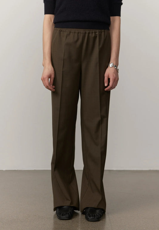A model is shown from the chest down wearing dark brown wide-leg trousers, a black top, and black shoes, standing against a neutral light gray wall.