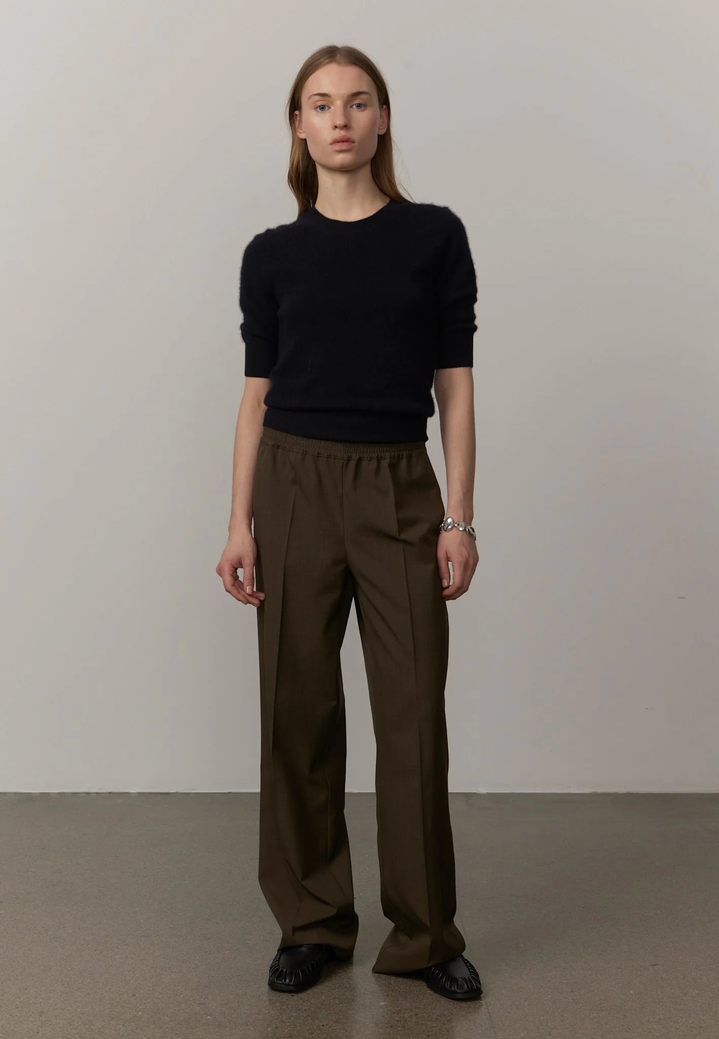 A woman stands against a plain light gray wall wearing a black short-sleeve crewneck sweater and dark olive brown wide-leg trousers, with black shoes.