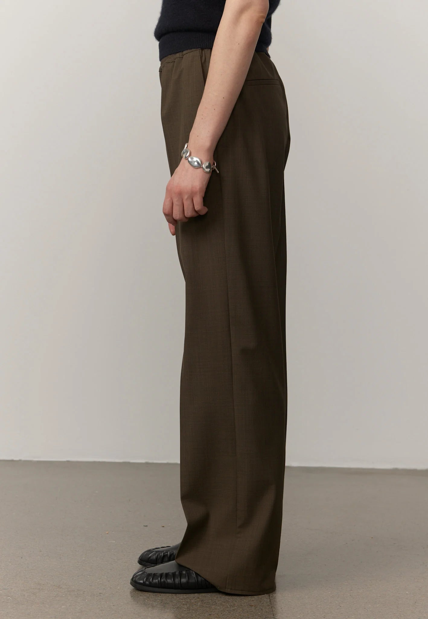 A person shown from the waist down wearing dark olive brown wide-leg trousers, a dark navy top, and black loafers, standing against a plain light gray wall.