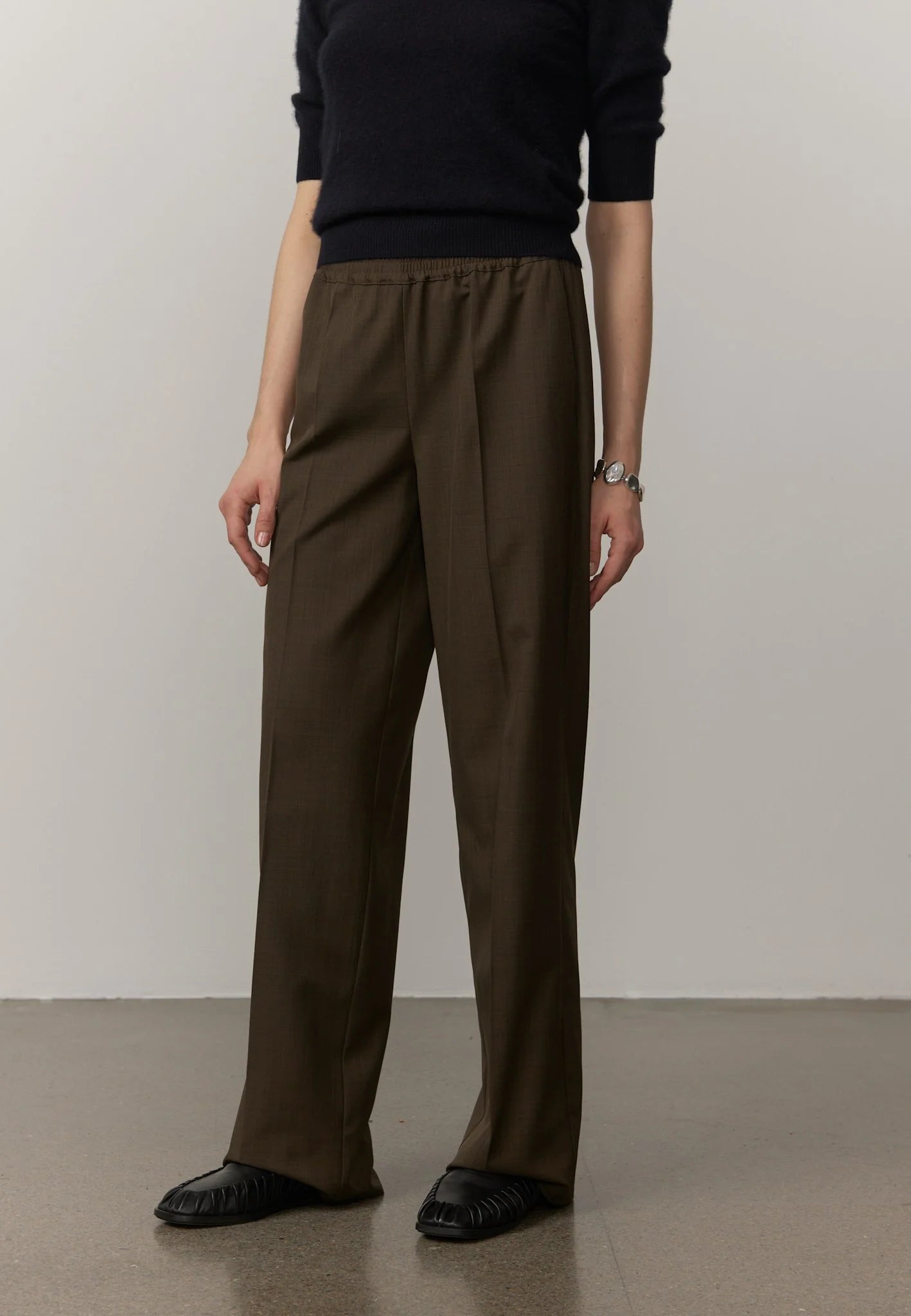 A model wearing dark brown wide-leg trousers with a drawstring waist, paired with a black long-sleeve top and black leather shoes, standing against a neutral studio background.
