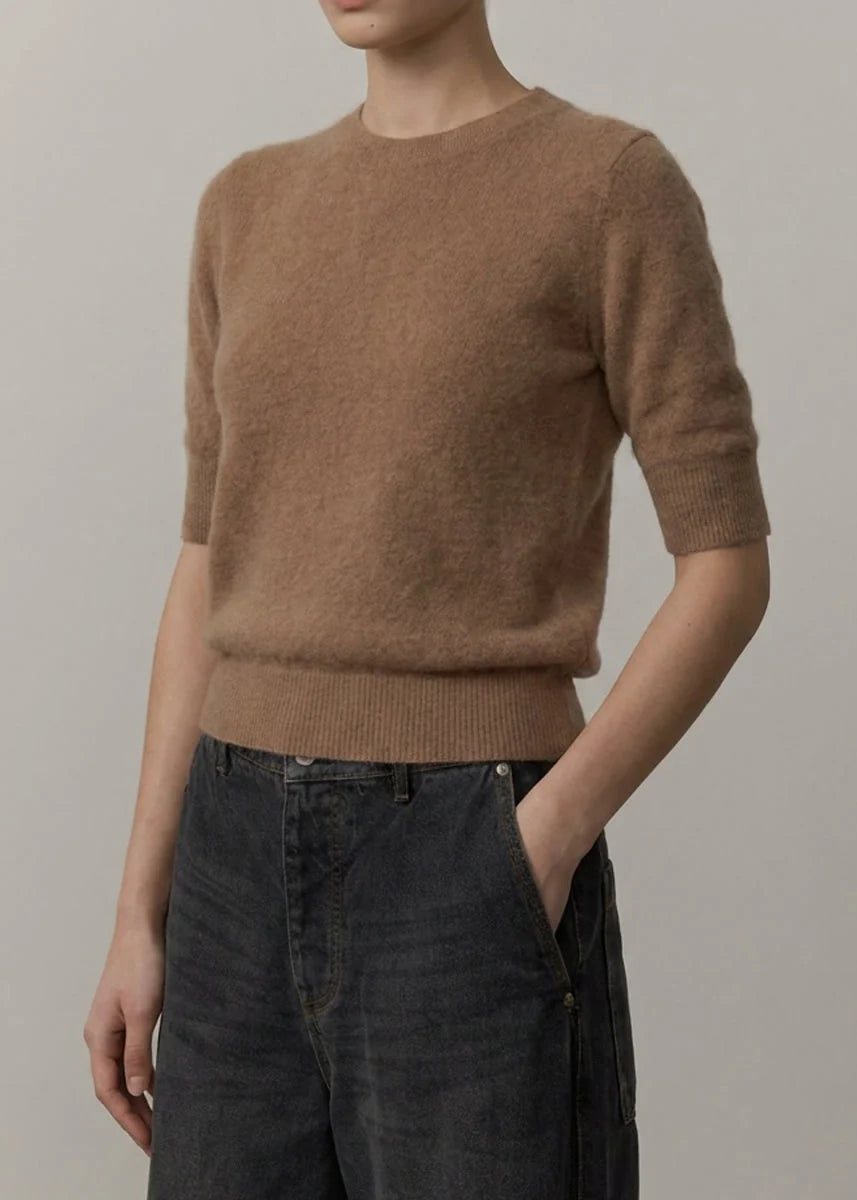 Close-up of a person wearing a soft camel brown knit sweater with short sleeves and a ribbed hem, paired with black bottoms.