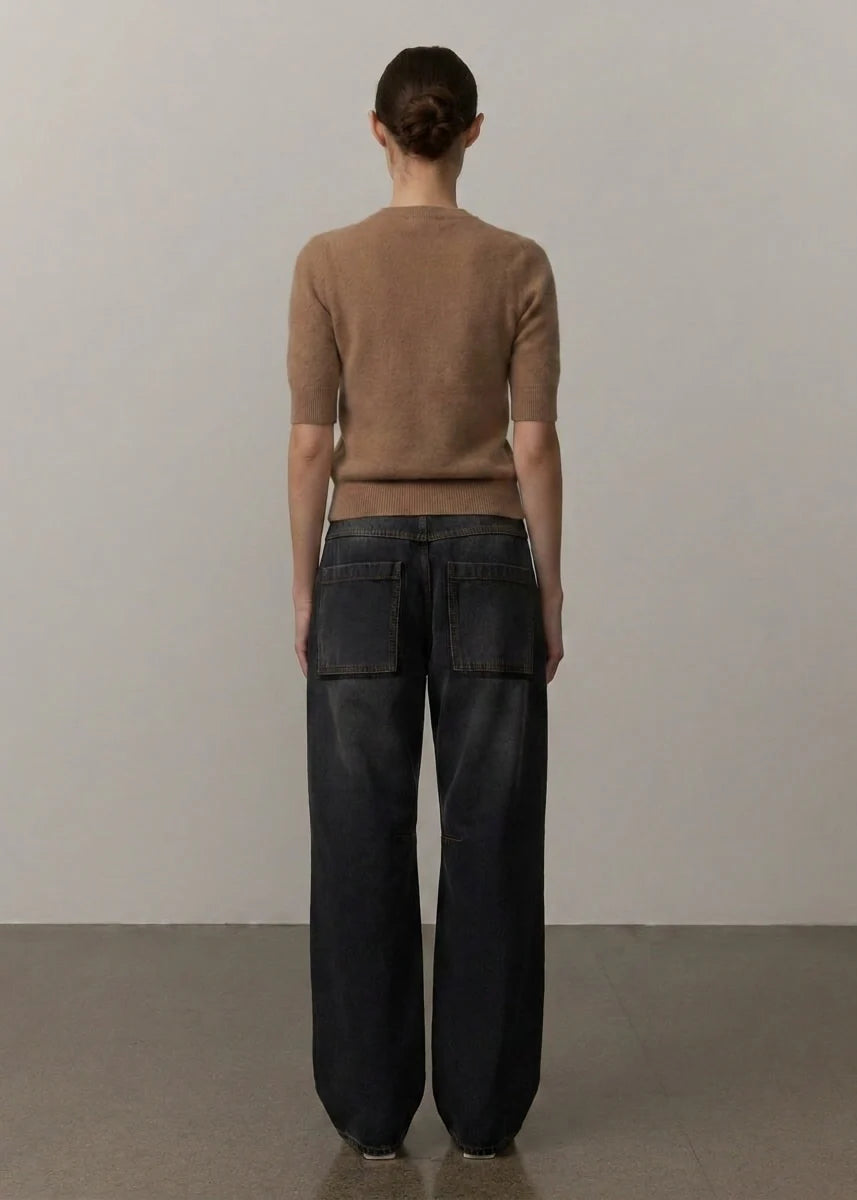 A model viewed from behind wearing a short-sleeve tan knit sweater and dark wide-leg jeans with back patch pockets, standing against a plain light gray wall.