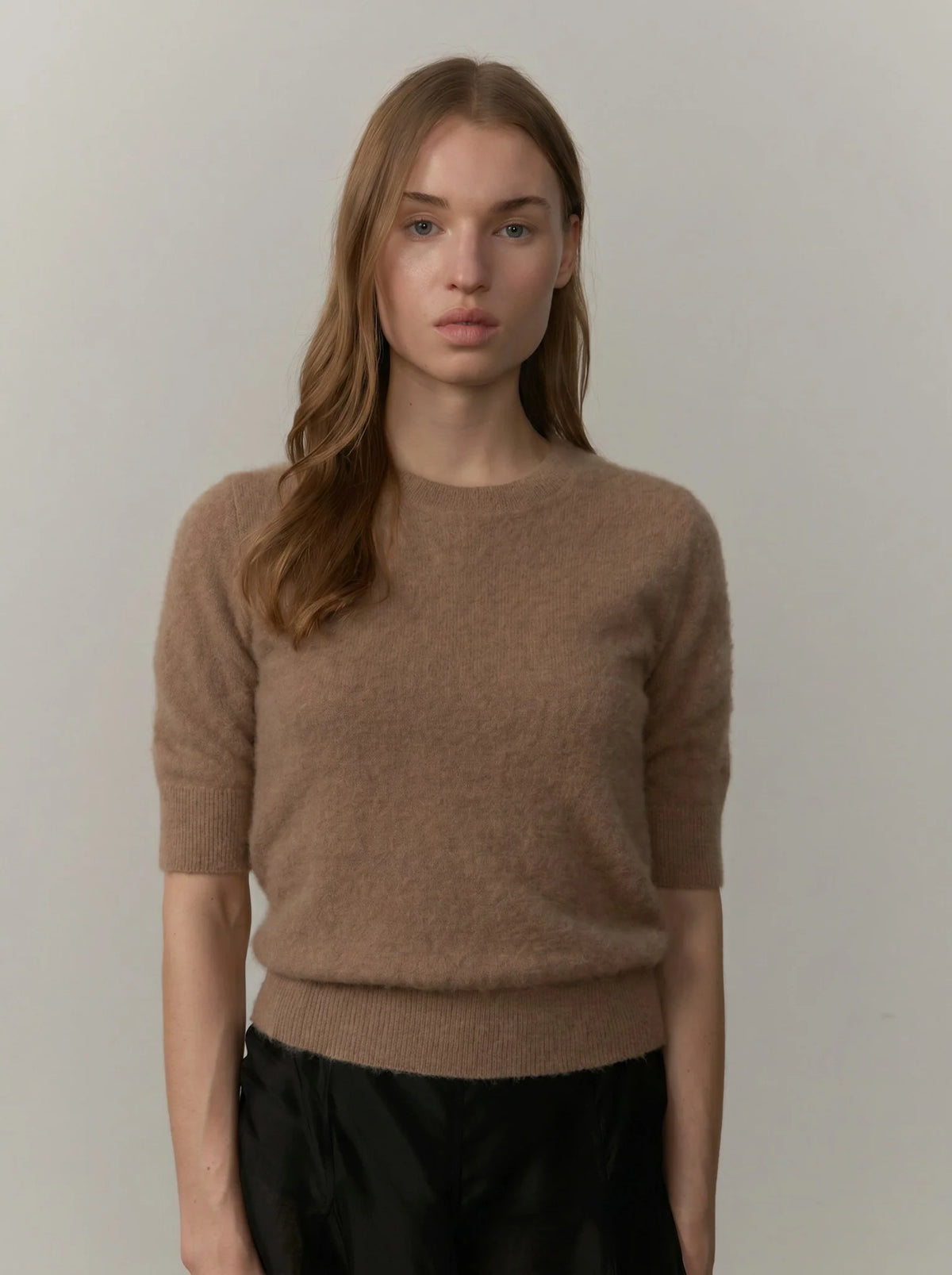 A woman wearing a short-sleeve crewneck knit sweater in a warm taupe-brown color, paired with black bottoms, standing against a plain light gray studio background.