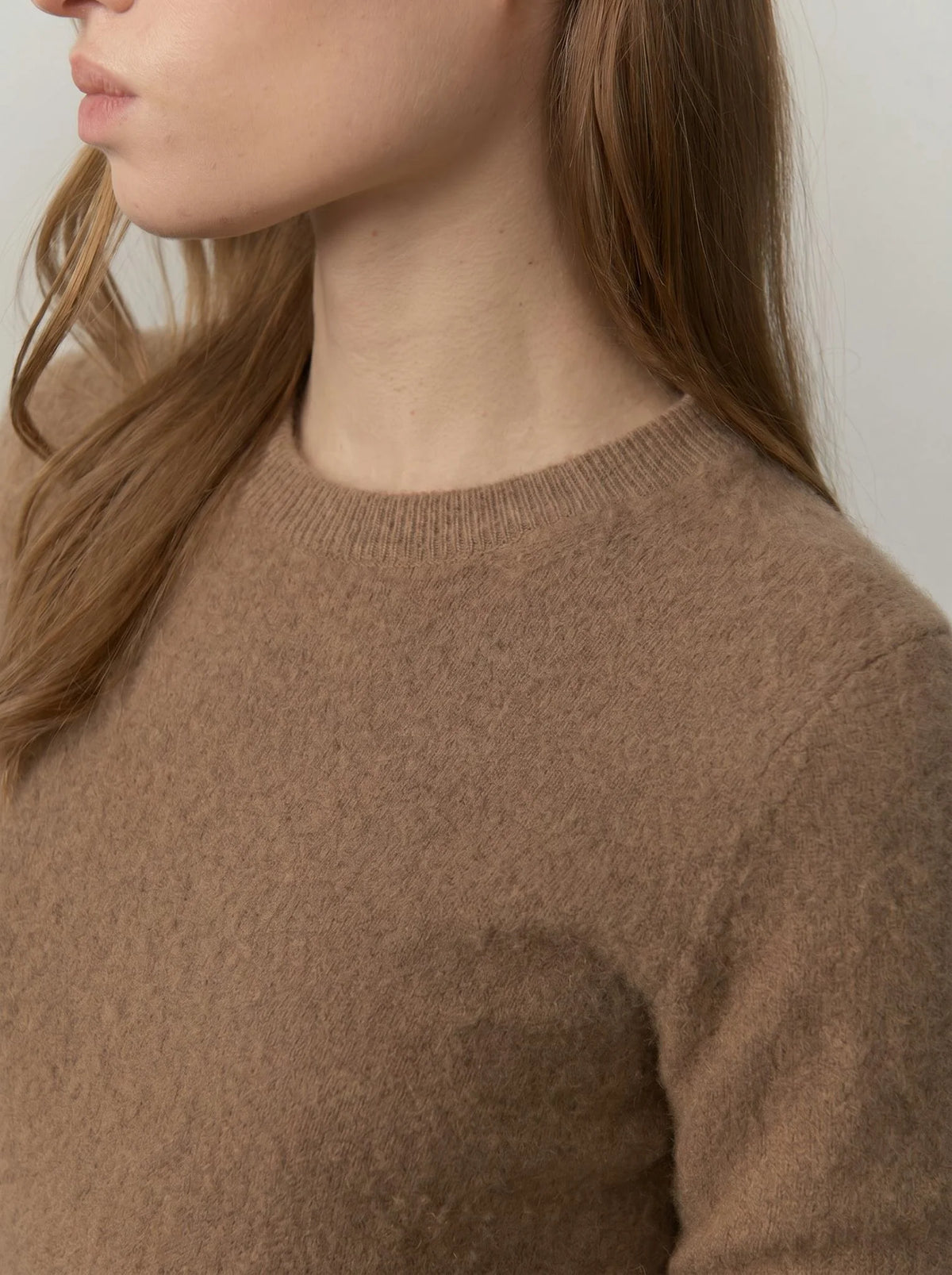 Close-up of a person wearing a soft knit crew-neck sweater in a warm taupe-brown color, with long light brown hair visible against a plain light background.
