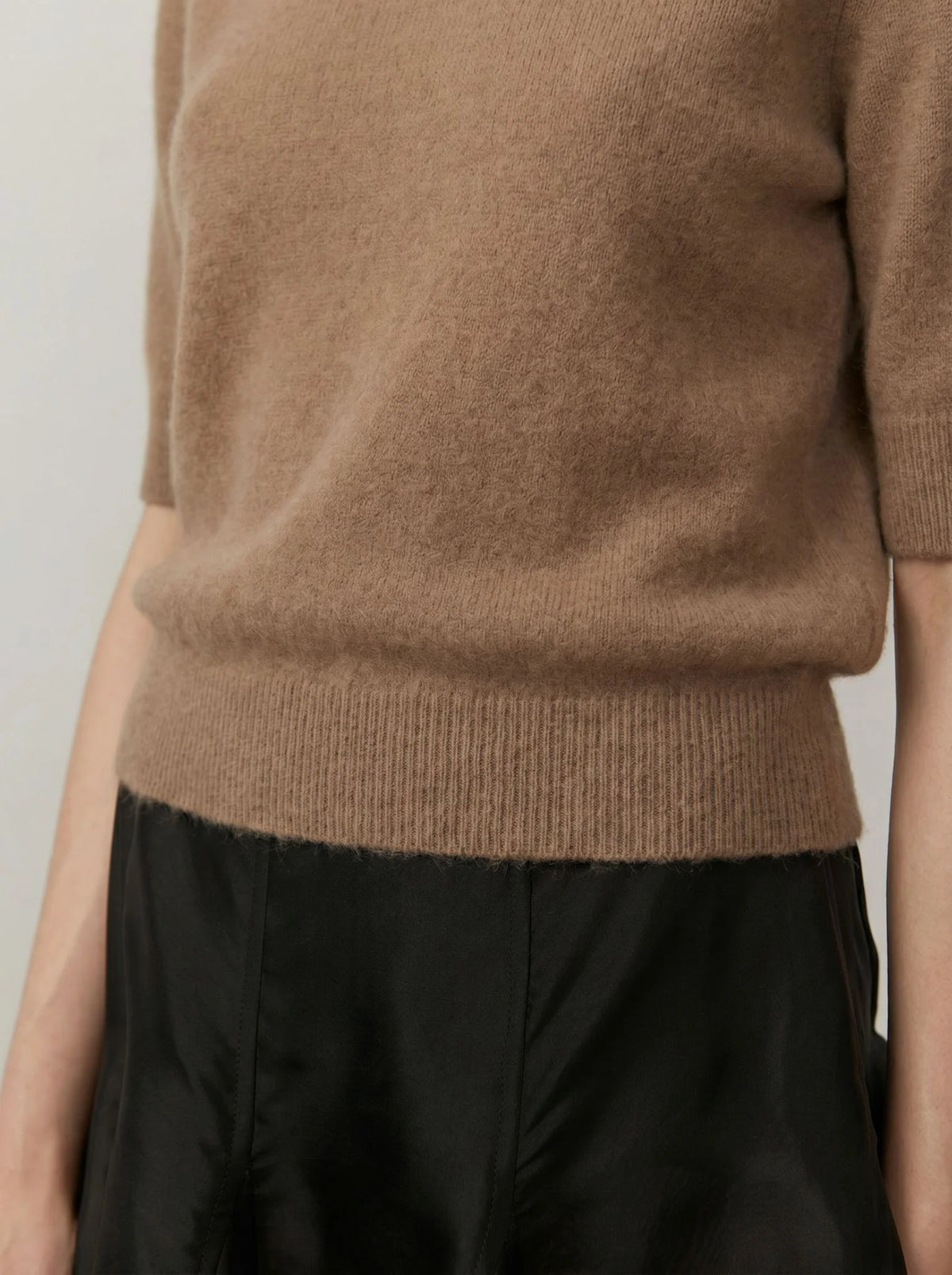 Close-up of a person wearing a soft camel brown knit sweater with short sleeves and a ribbed hem, paired with black bottoms.