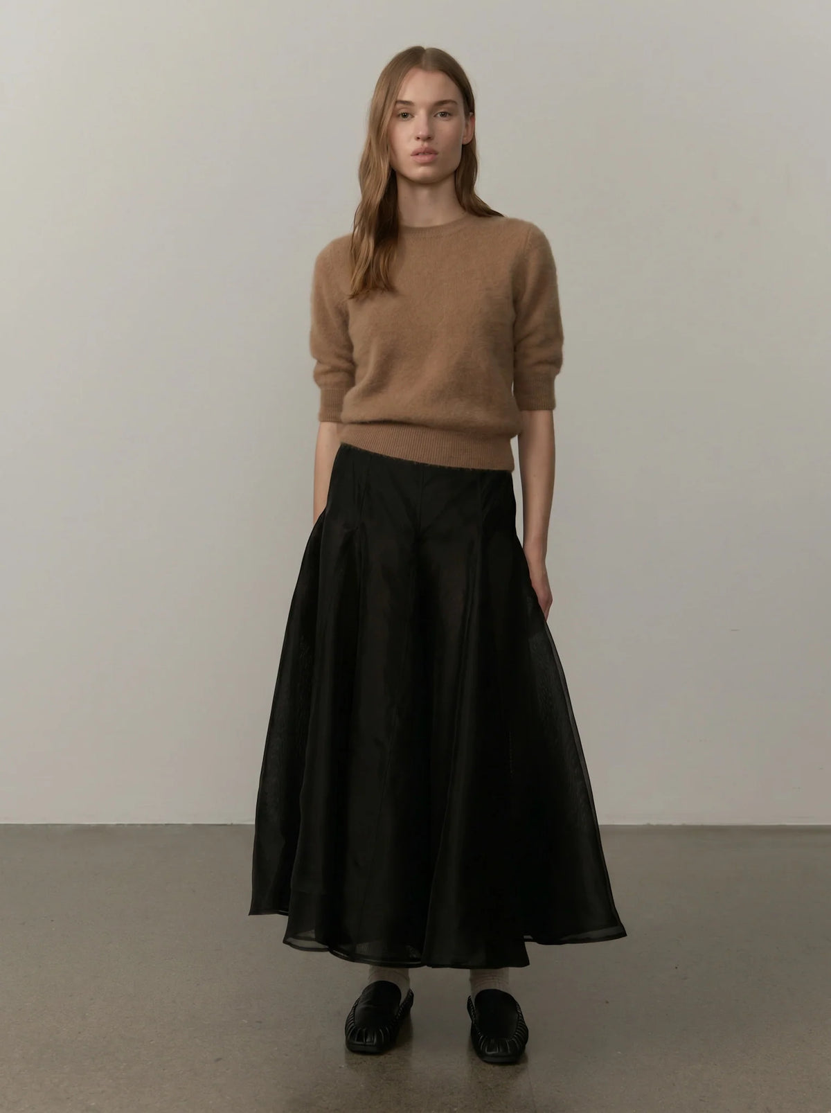 A woman stands in a studio wearing a tan knit sweater and a long black skirt, paired with black shoes.