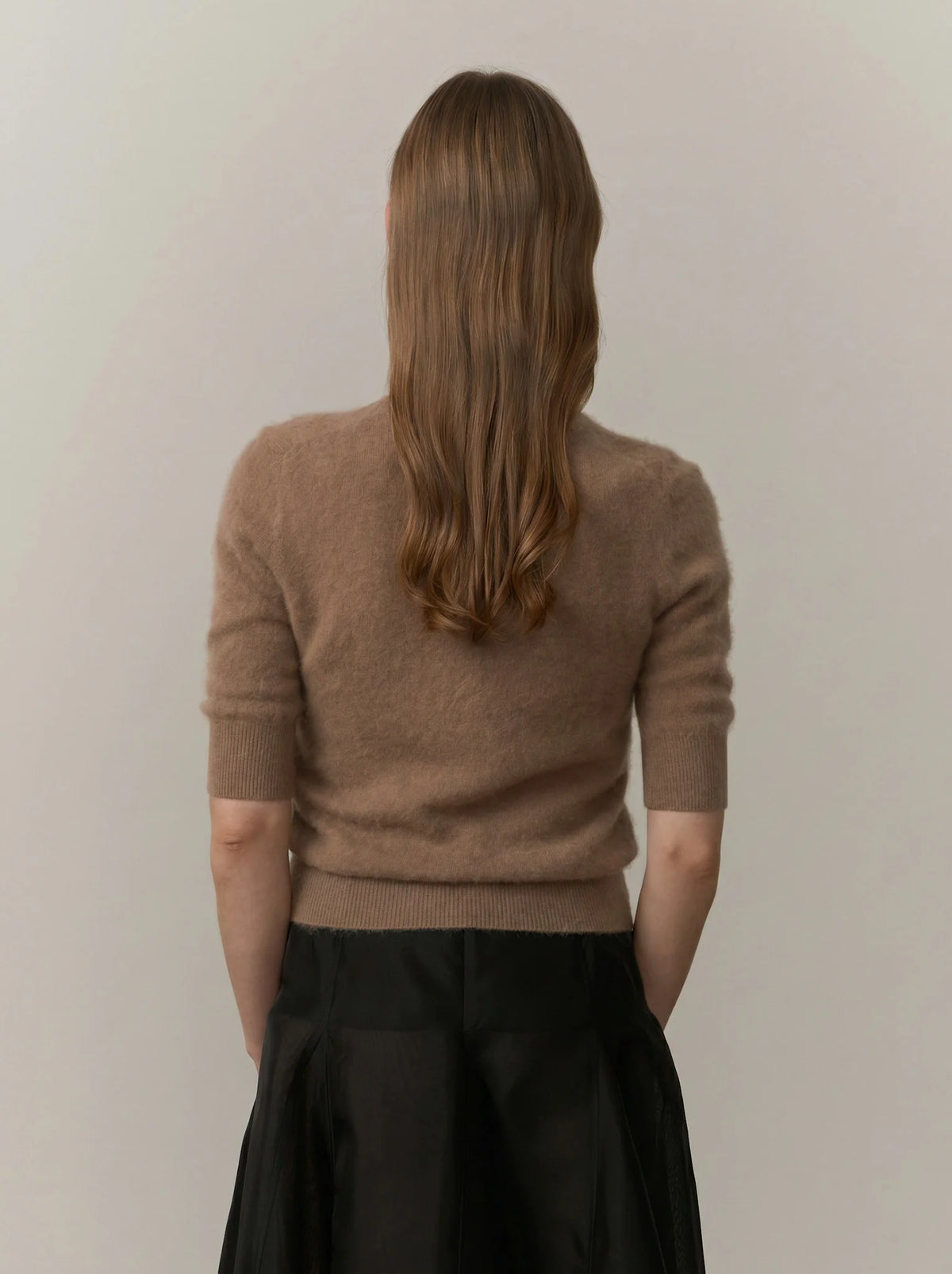 A woman viewed from behind wearing a short-sleeve taupe knit sweater and a black skirt, standing against a plain light gray studio background.