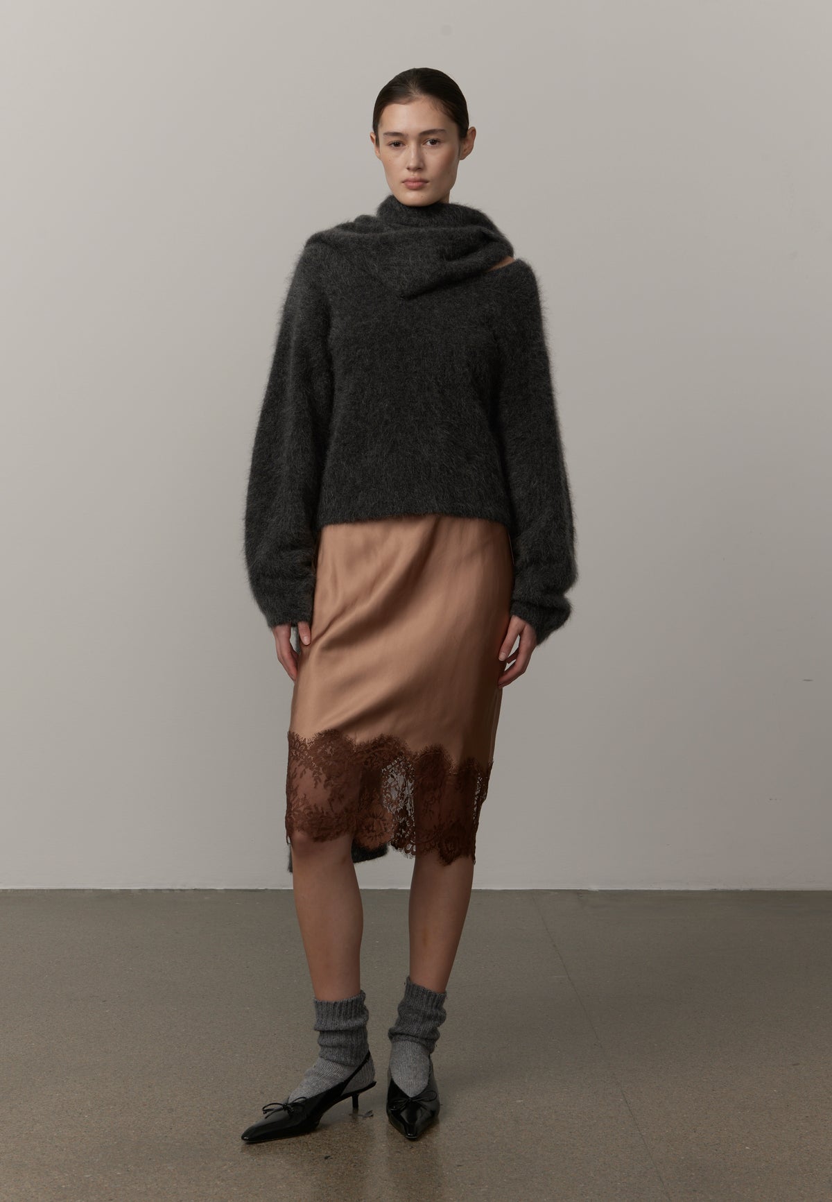 A model stands against a plain light gray studio wall wearing a dark charcoal fuzzy cropped sweater, a tan satin midi skirt with a brown lace hem, gray socks, and black pointed heels.