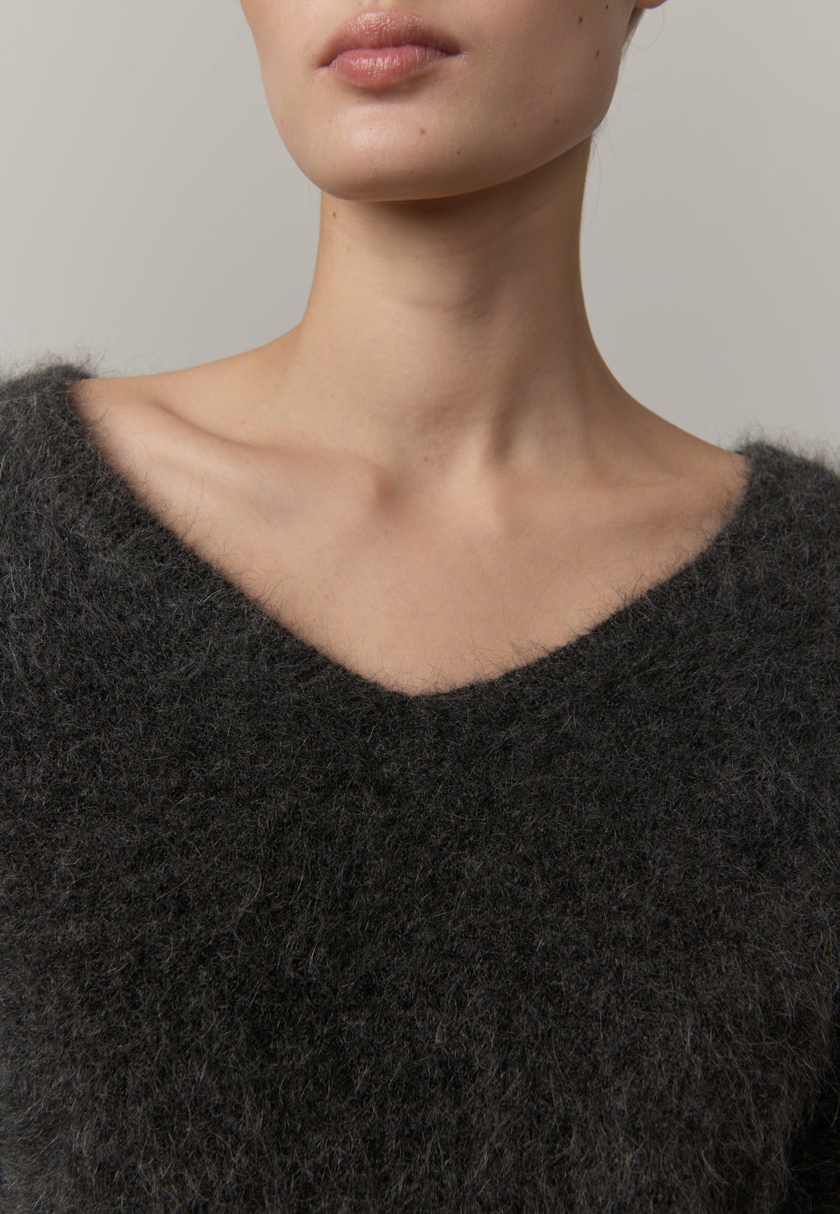 Close-up of a model wearing a dark charcoal gray fuzzy V-neck sweater against a neutral light gray background.
