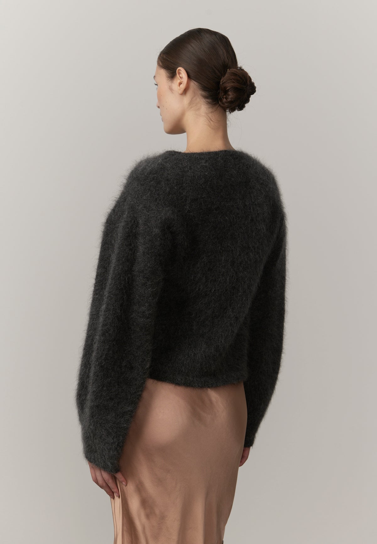 A woman shown from the back wearing a dark charcoal fuzzy cropped sweater and a light beige satin skirt, set against a plain light gray studio background.
