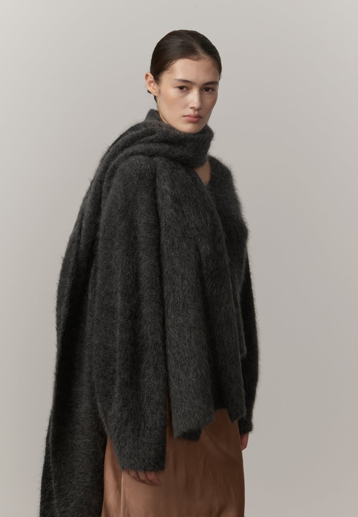 A model wearing a dark gray fuzzy, oversized wrap or poncho-style sweater with a high folded collar, paired with brown bottoms, against a plain light gray studio background.